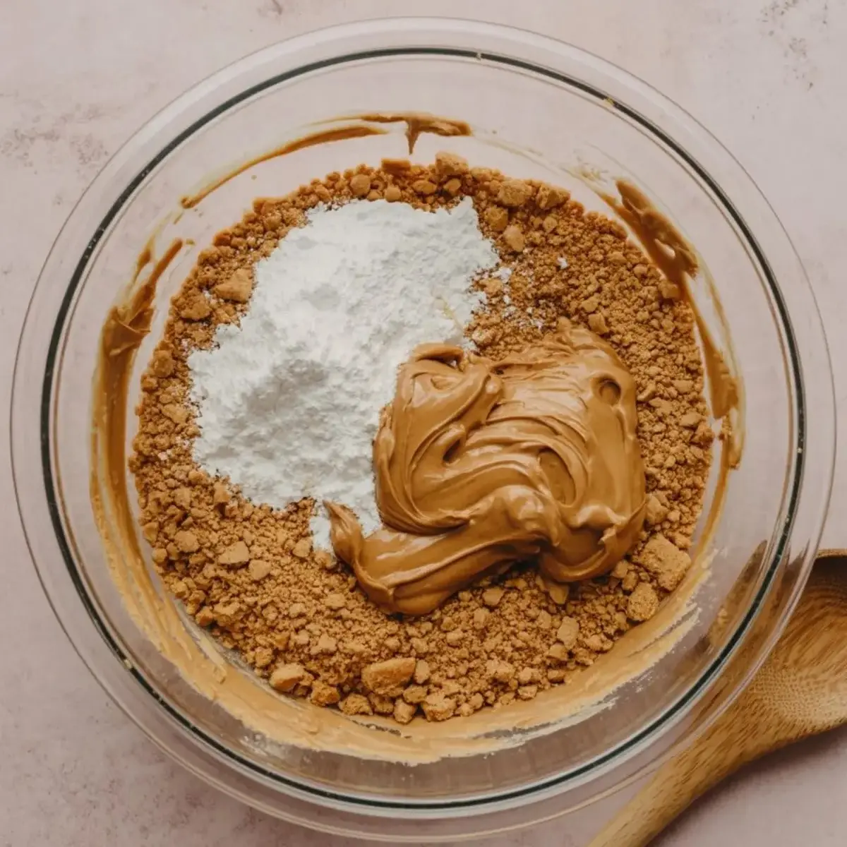 Mixing bowl filled with graham cracker crumbs, powdered sugar, and creamy peanut butter on a pink surface with a wooden spoon beside it.