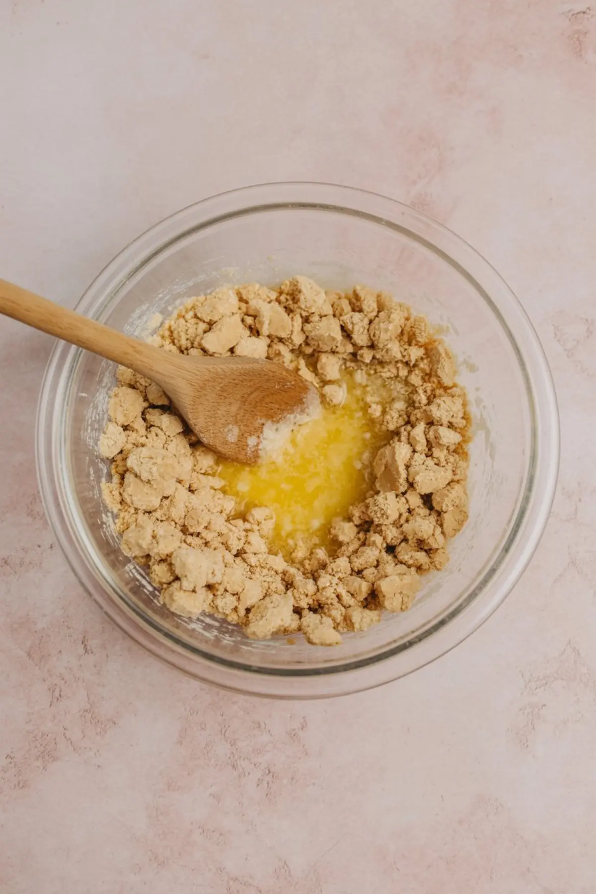 Glass mixing bowl with a wooden spoon, showing partially combined crushed graham crackers and melted butter for a no-bake dessert crust on a light pink surface.