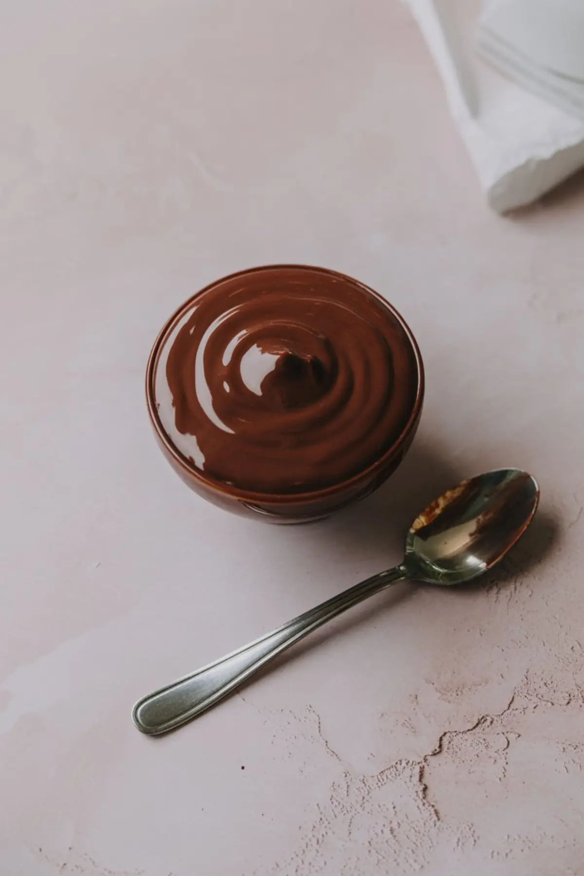Small bowl of glossy melted chocolate with swirls on top, next to a spoon with chocolate residue, placed on a soft pastel surface.