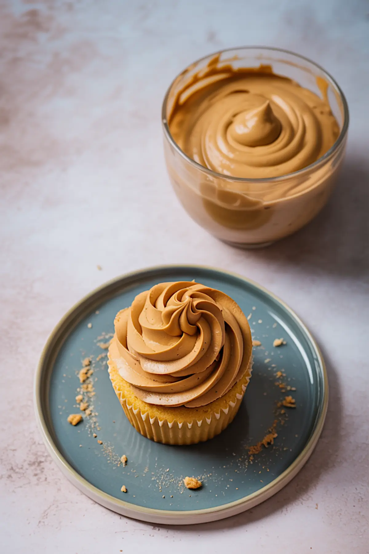 Glass bowl of swirled peanut butter frosting placed next to a frosted cupcake on a teal plate, with scattered crumbs on a light surface.