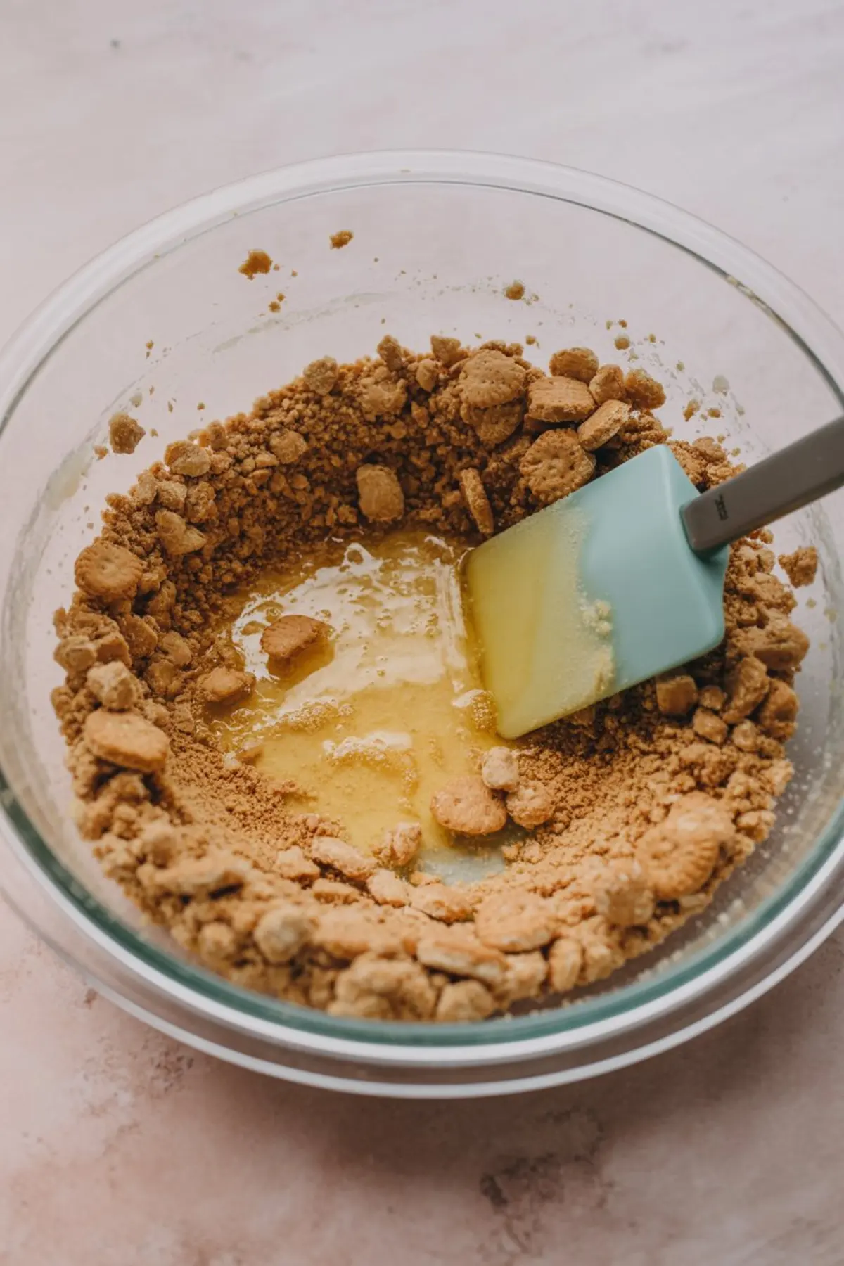  A glass mixing bowl filled with crushed cookies and melted butter being combined with a spatula on a light pink surface.