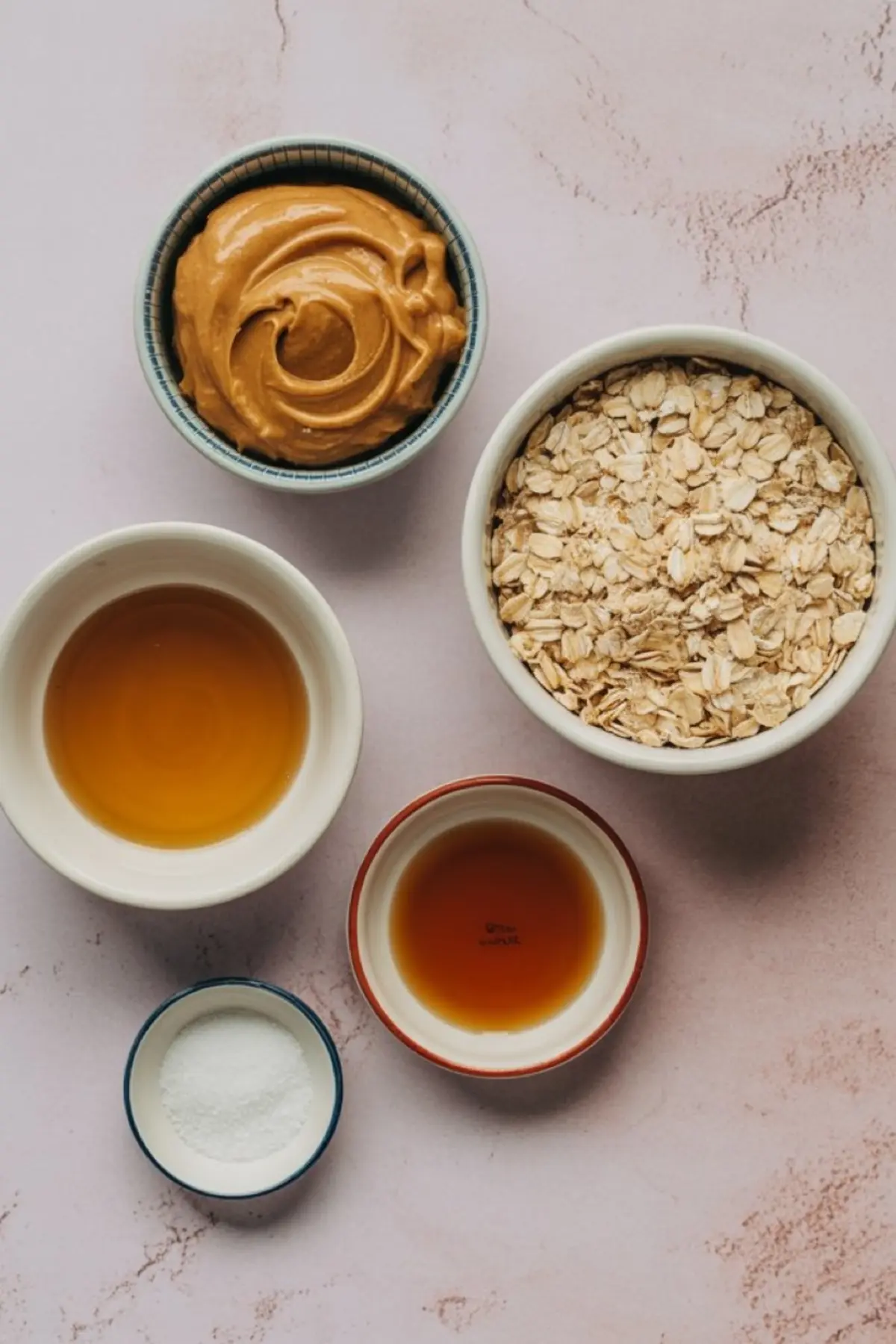Overhead image of five small bowls on a pink surface filled with ingredients for no-bake snacks, including creamy peanut butter, rolled oats, maple syrup, vanilla extract, and salt.
