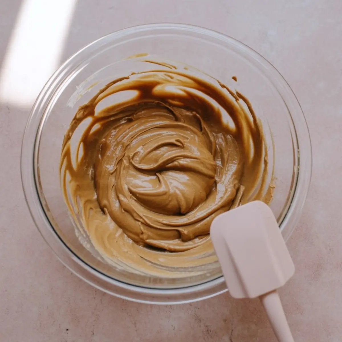 Glass mixing bowl with a smooth blend of peanut butter and maple syrup being stirred with a silicone spatula, shown on a pink background.