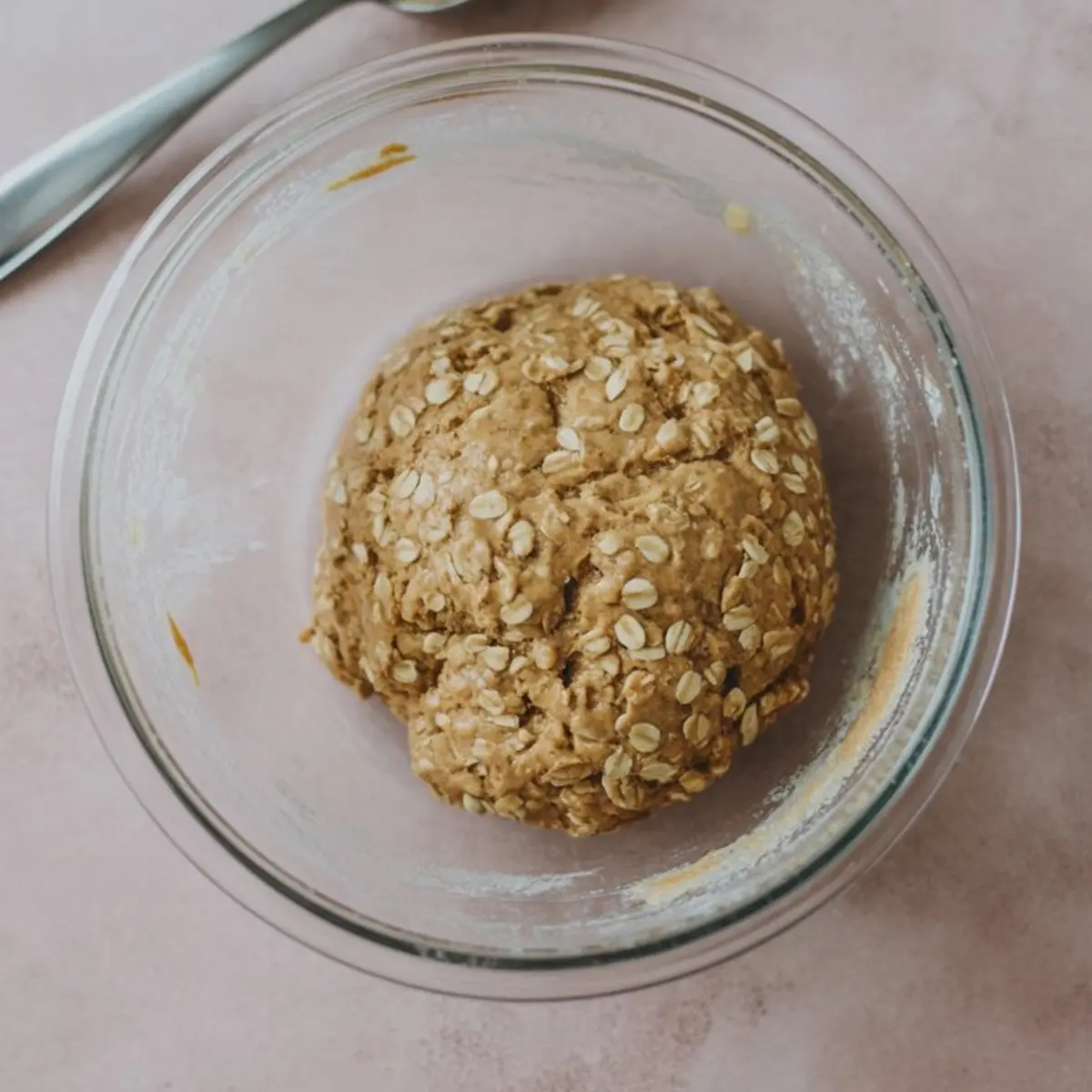 Dough mixture of oats and peanut butter rolled into a soft ball in a clear glass bowl, placed on a pink surface.