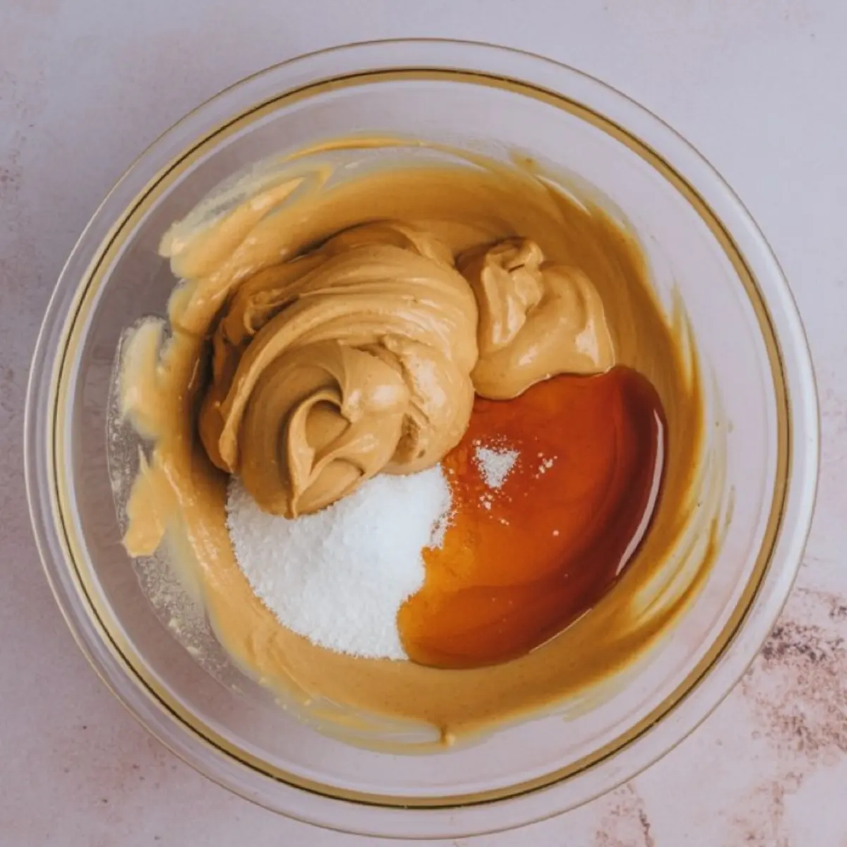 Peanut butter, maple syrup, and sugar combined in a glass bowl before mixing, ingredients laid out for homemade protein snack prep.