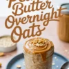 Jar of peanut butter overnight oats topped with creamy peanut butter and crushed peanuts, styled on a blue plate with scattered oats and a spoon on a pink surface.