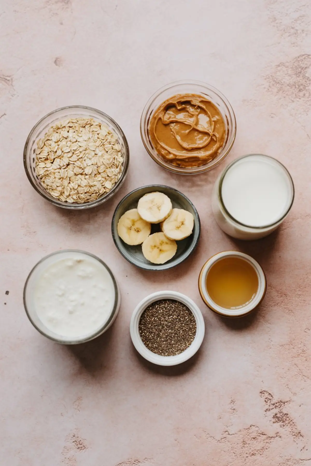 Flat lay of peanut butter overnight oats ingredients including rolled oats, peanut butter, banana slices, milk, yogurt, chia seeds, and maple syrup in glass bowls on a light pink background.
