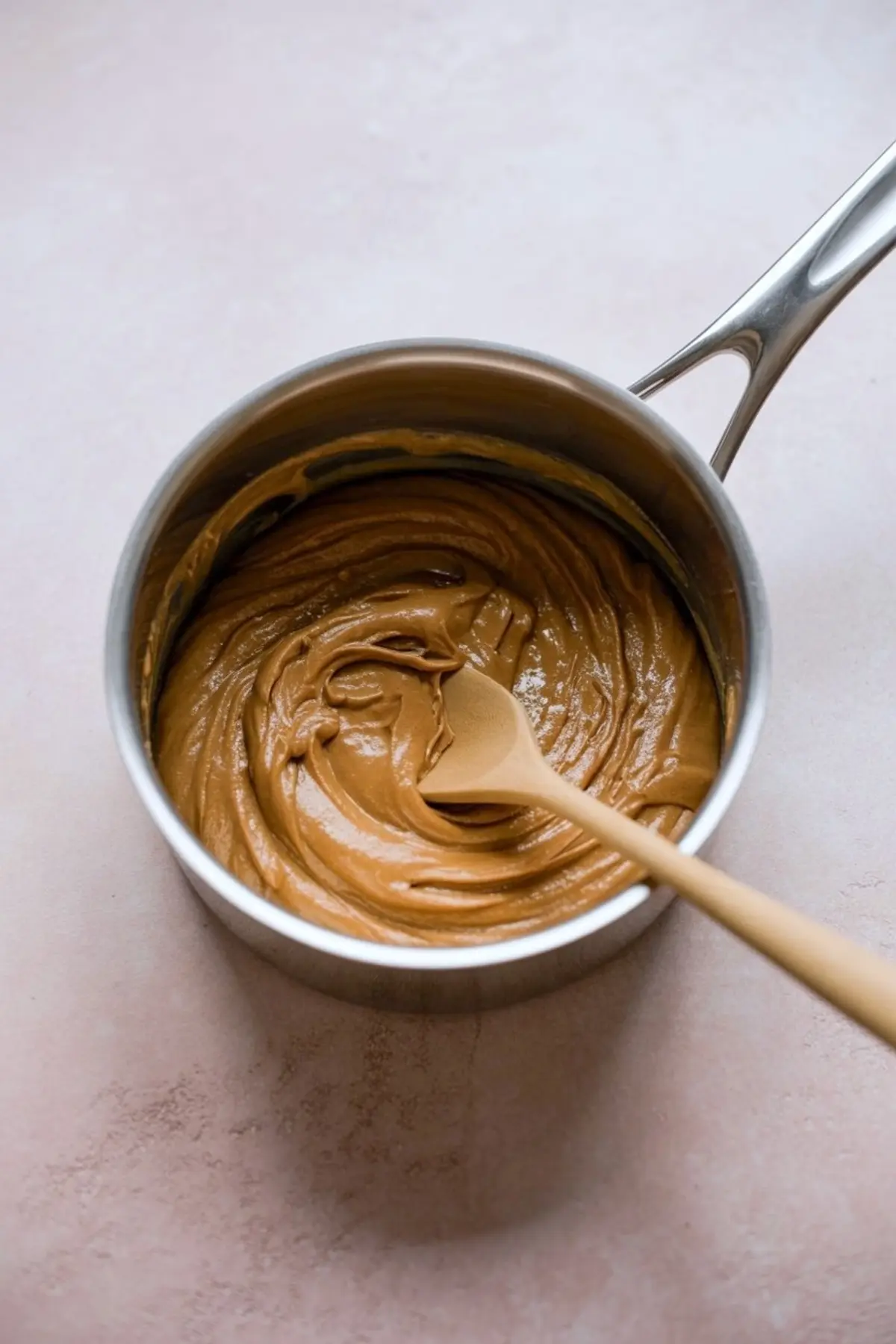 Peanut butter mixture melted in a saucepan and swirled with a wooden spoon on a light countertop.