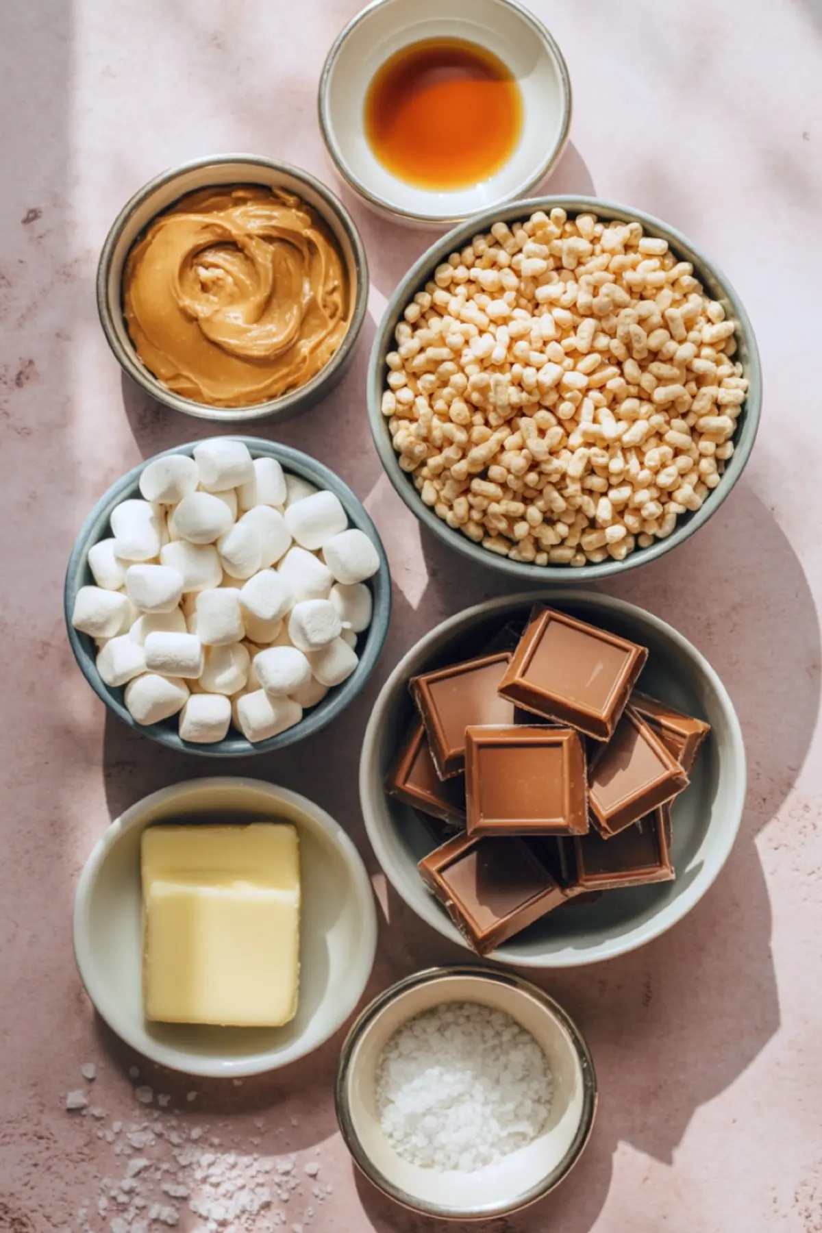 Flat lay of peanut butter Rice Krispie treat ingredients on a pink surface, including mini marshmallows, peanut butter, vanilla extract, puffed rice cereal, butter, milk chocolate squares, and flaky sea salt.
