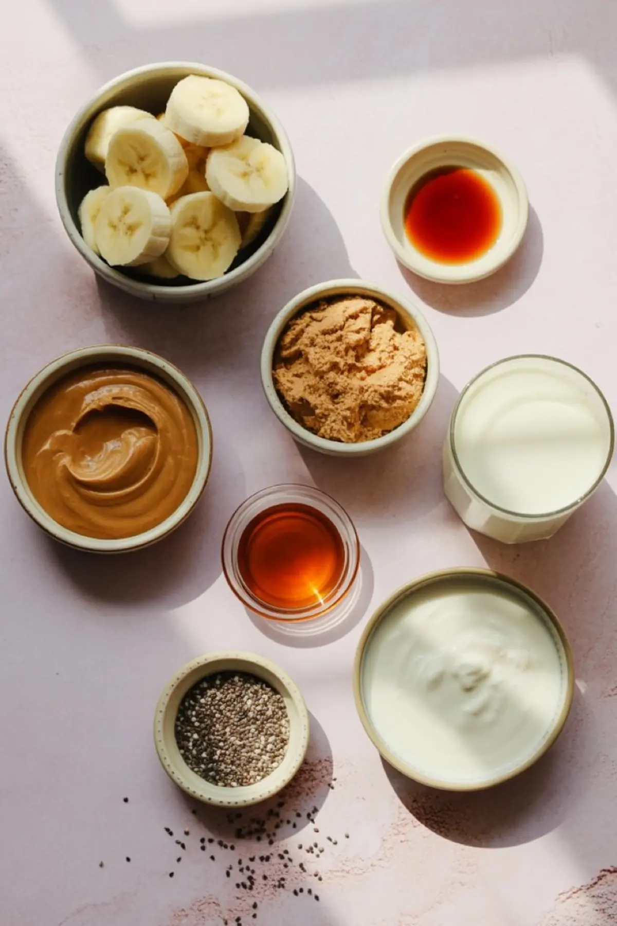 Ingredients for a peanut butter banana smoothie including sliced bananas, peanut butter, protein powder, chia seeds, yogurt, milk, vanilla extract, and honey arranged in small bowls on a light surface.