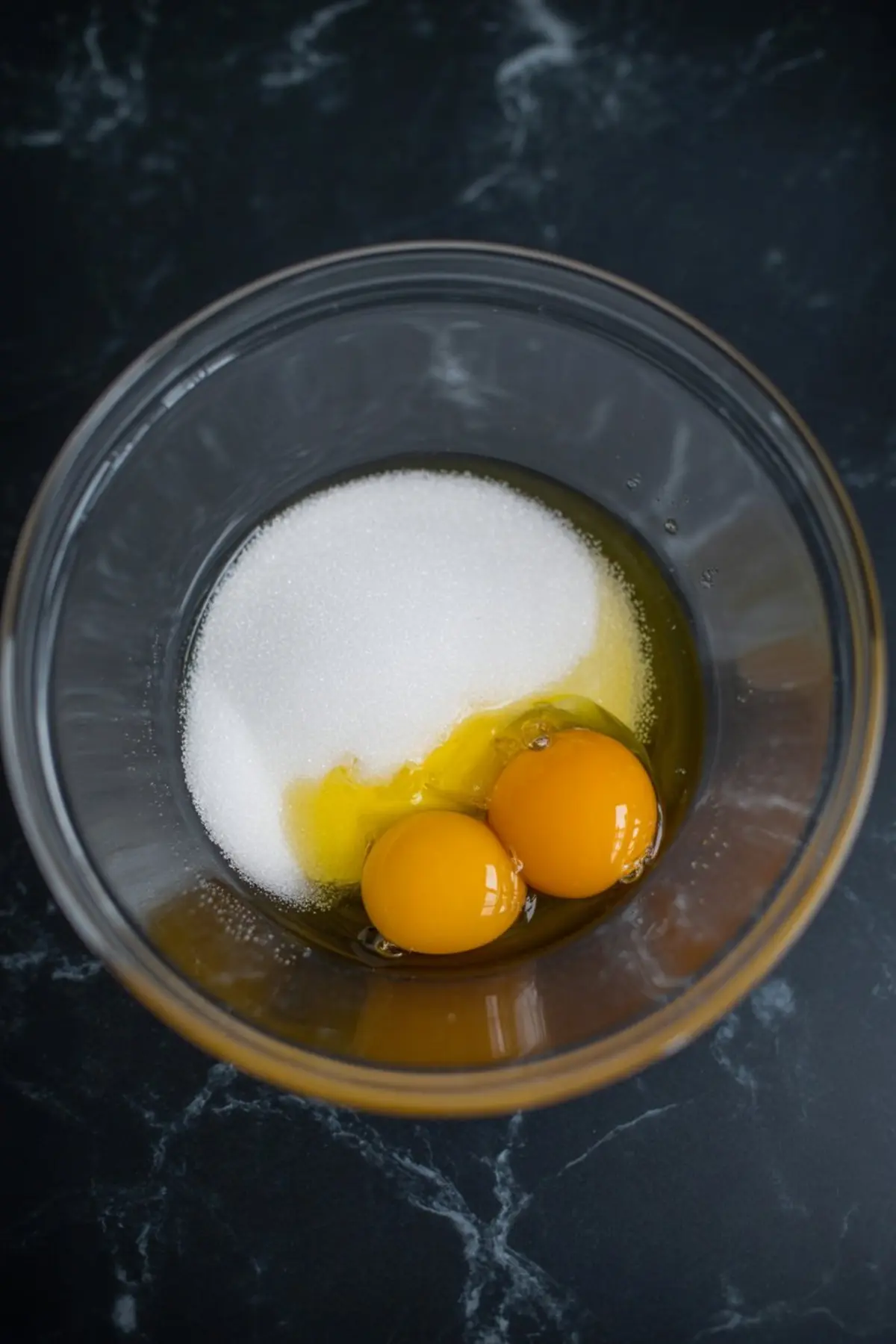 Close-up of a glass bowl with two raw eggs, granulated sugar, and oil, ready for mixing on a dark stone surface.