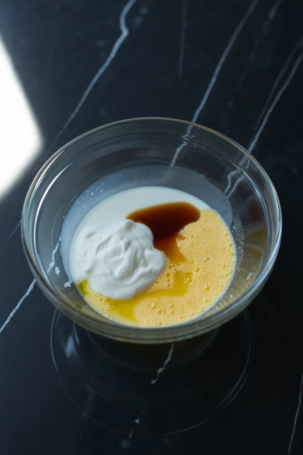 Mixing bowl with wet ingredients including yogurt, eggs, milk, and vanilla extract on a black marble background.