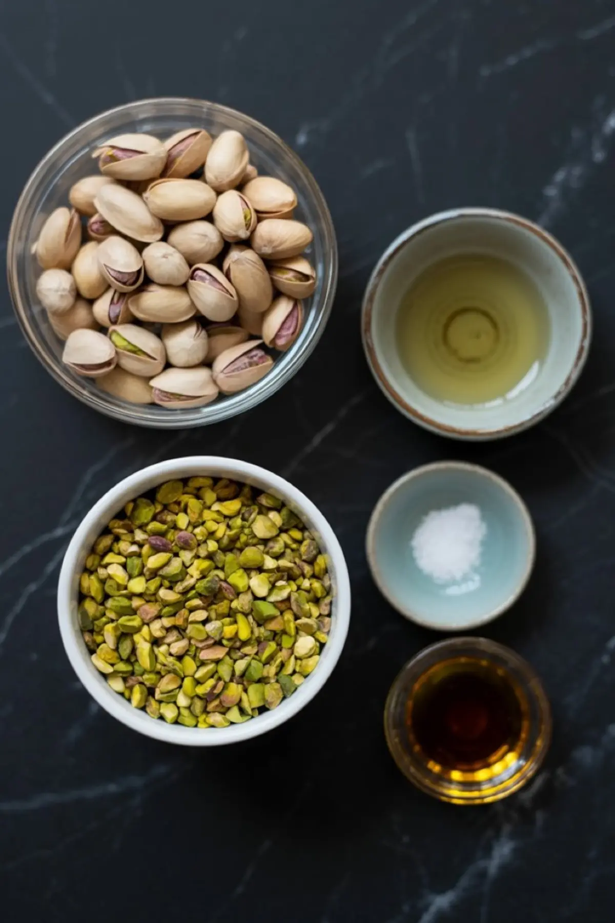 Flat lay of pistachio butter ingredients including whole pistachios, chopped pistachios, oil, salt, and maple syrup arranged in ceramic bowls on a dark marble surface.