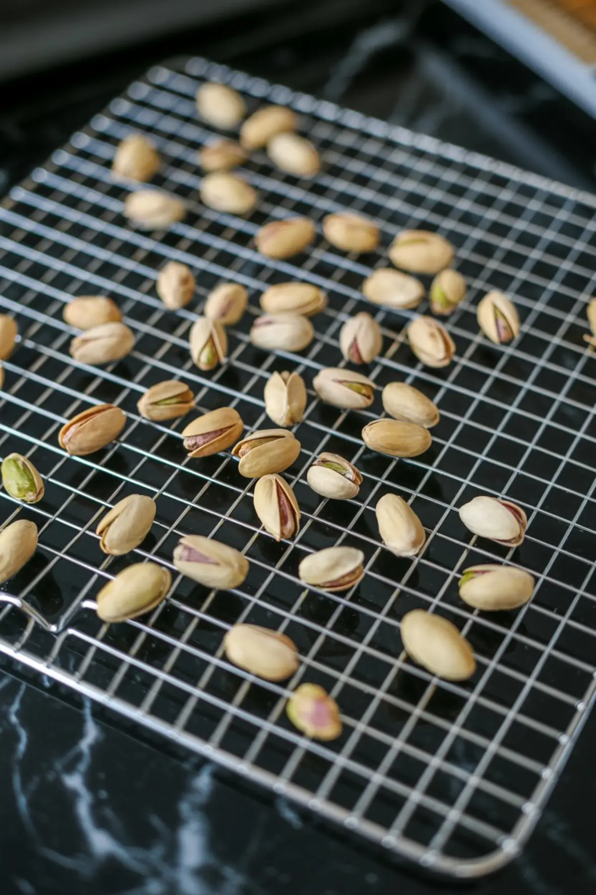 Roasted pistachios cooling on a metal wire rack, showcasing open shells and toasted textures against a dark granite background.