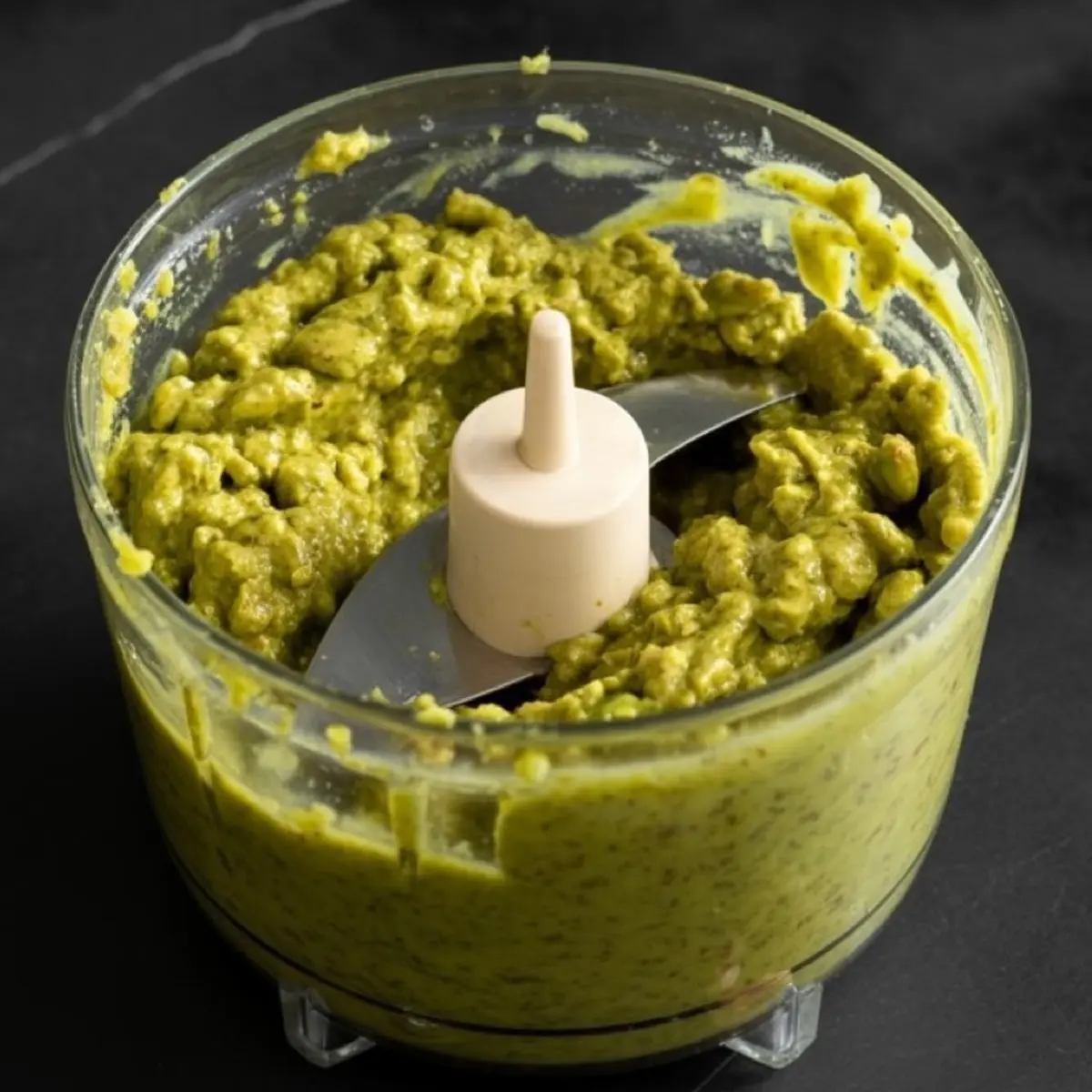 Blended pistachio butter in a food processor with a creamy, chunky texture and vibrant green color, freshly mixed on a black surface.