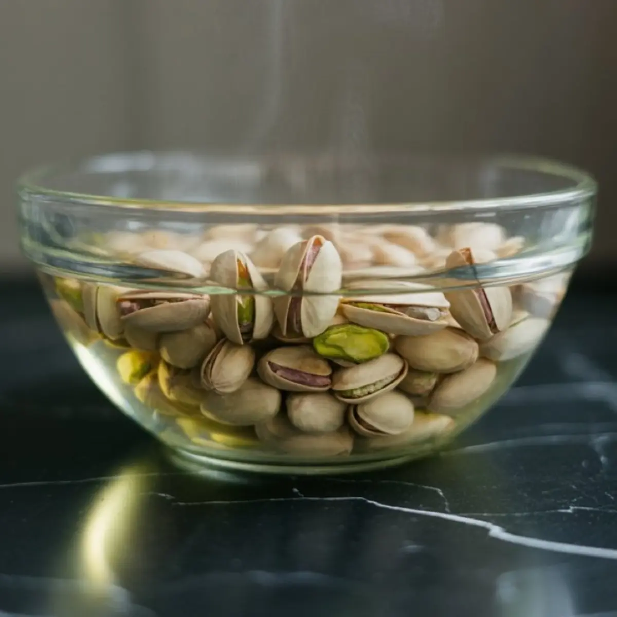 Glass bowl filled with whole pistachios soaking in water on a dark marble countertop, ready for shelling and baking preparation.