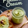 Homemade pistachio cream in a glass jar with a smooth, glossy texture, served on toasted bread and garnished with crushed pistachios, styled with a gold spoon on a pink plate.