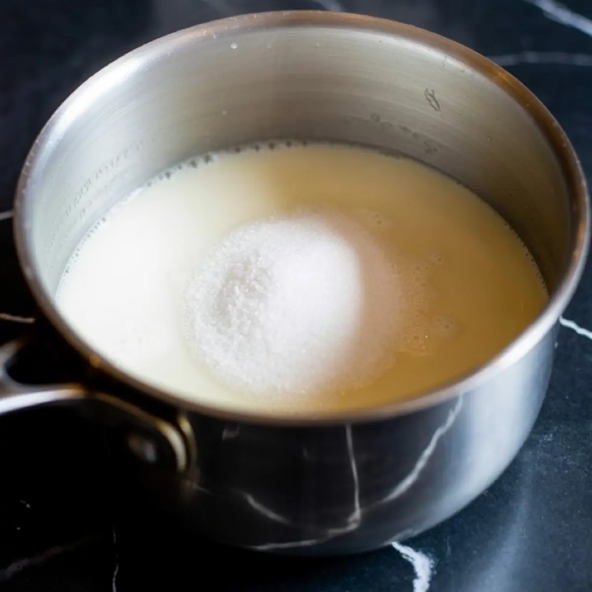 Stainless steel saucepan filled with warm milk and a mound of granulated sugar, placed on a black marble countertop during the early stages of making pistachio cream.