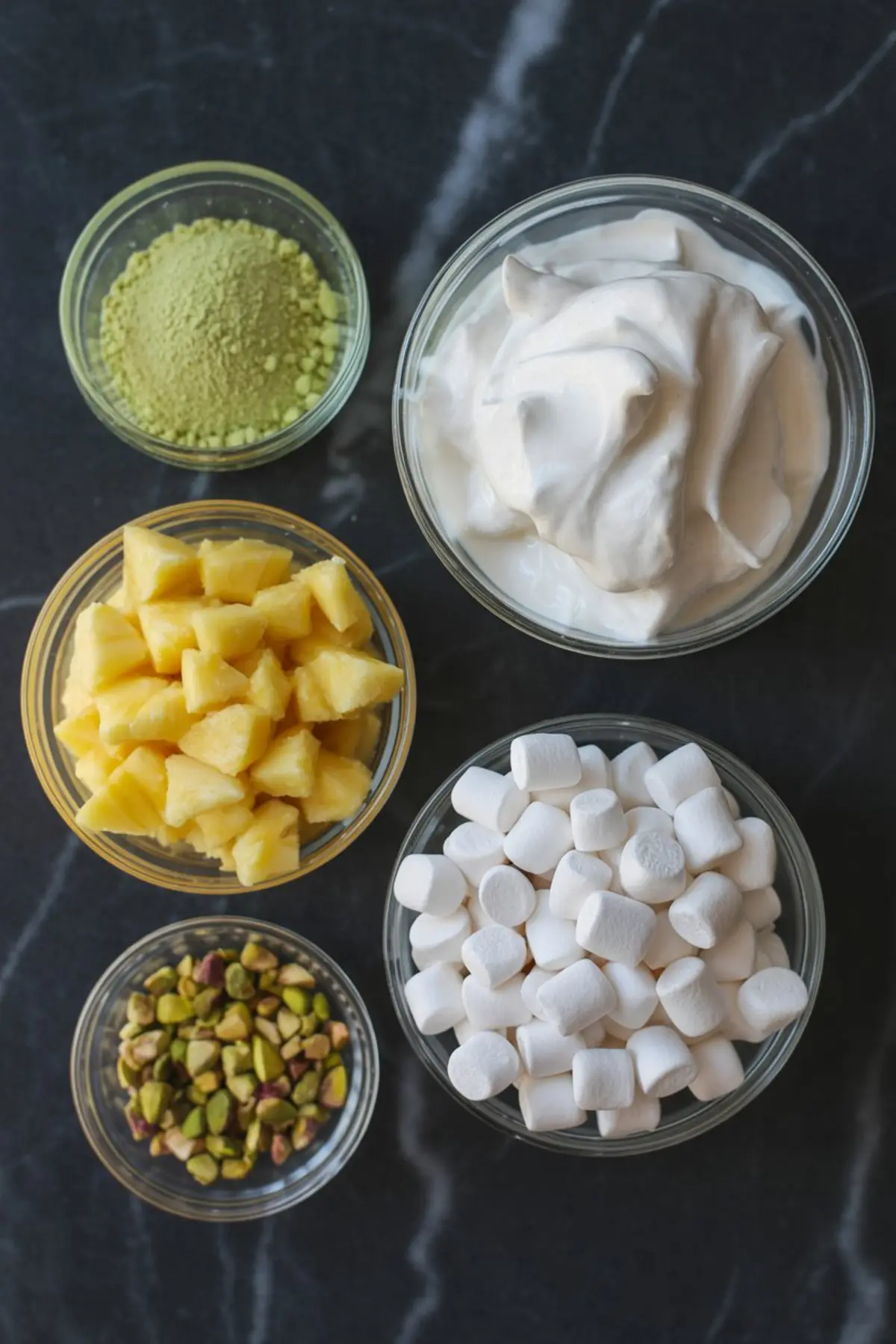 Flat lay of six clear bowls filled with ingredients for pistachio fluff salad, including pistachio pudding mix, whipped topping, pineapple chunks, mini marshmallows, chopped pistachios, and yogurt, arranged on a black marble surface.