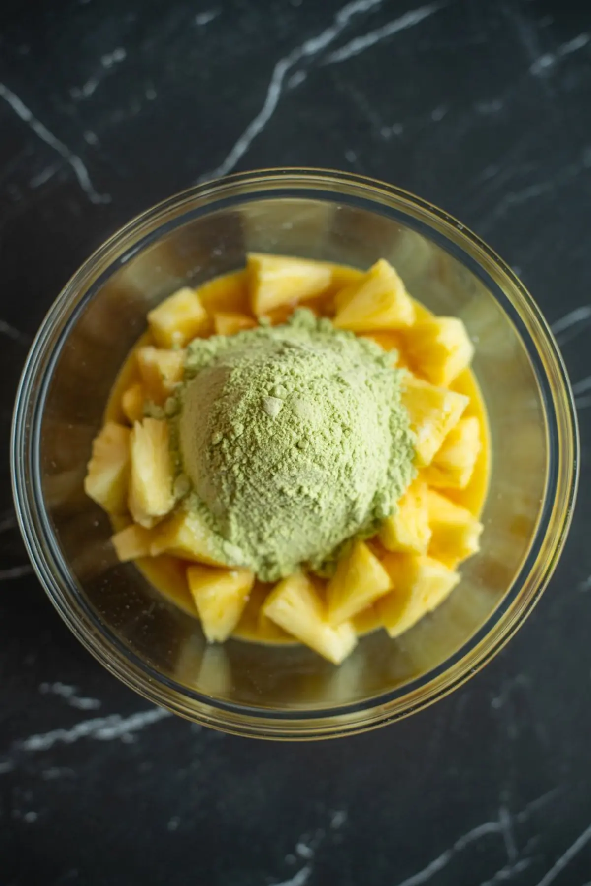 Overhead view of a glass bowl filled with pineapple chunks, orange juice, and a large heap of green pistachio pudding mix powder on top, sitting on a black marble countertop.