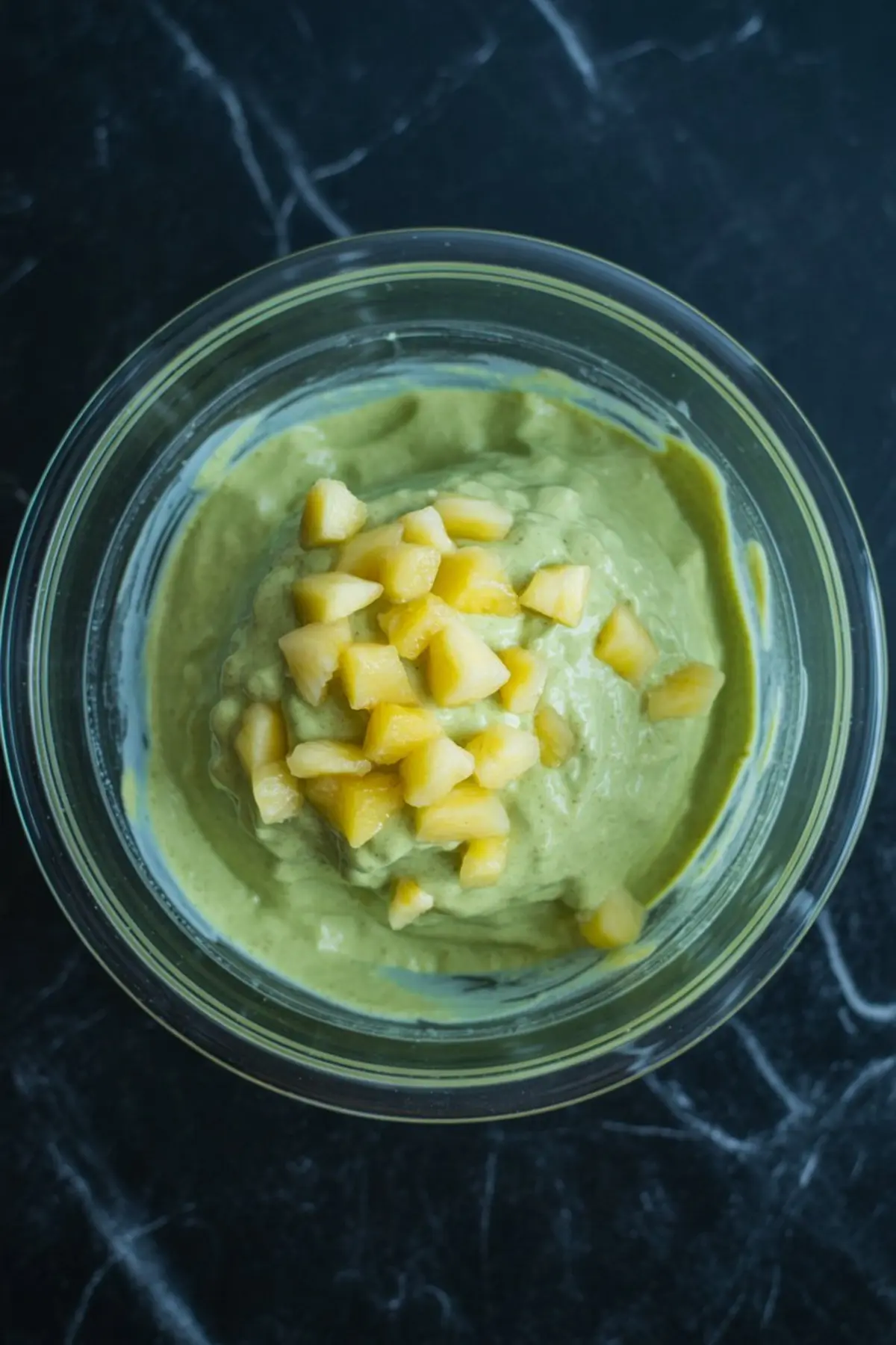 Smooth blended green pistachio pudding in a glass bowl, topped with finely diced pineapple pieces, photographed from above on a dark marble surface.
