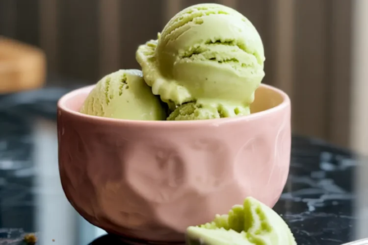 Close-up of pistachio ice cream scoops in a pink ceramic bowl, with a scoop on a spoon and scattered crushed pistachios on the glossy black surface, captioned “Pistachio Ice Cream.”