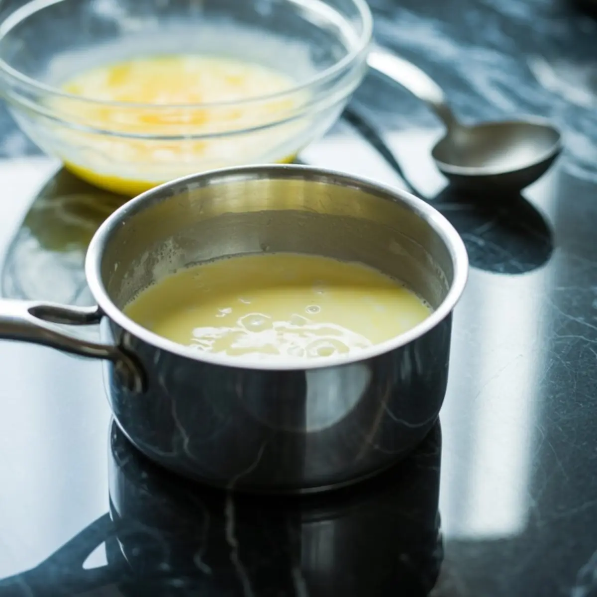 Stainless steel saucepan filled with a thick yellow custard mixture, set on a reflective black marble surface with a glass mixing bowl in the background.