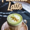 Pistachio latte in a clear glass mug with a swirl of green pistachio cream and white foam, topped with chopped nuts, set on a pink plate with a gold spoon and decorative “Pistachio Latte” text overlay.