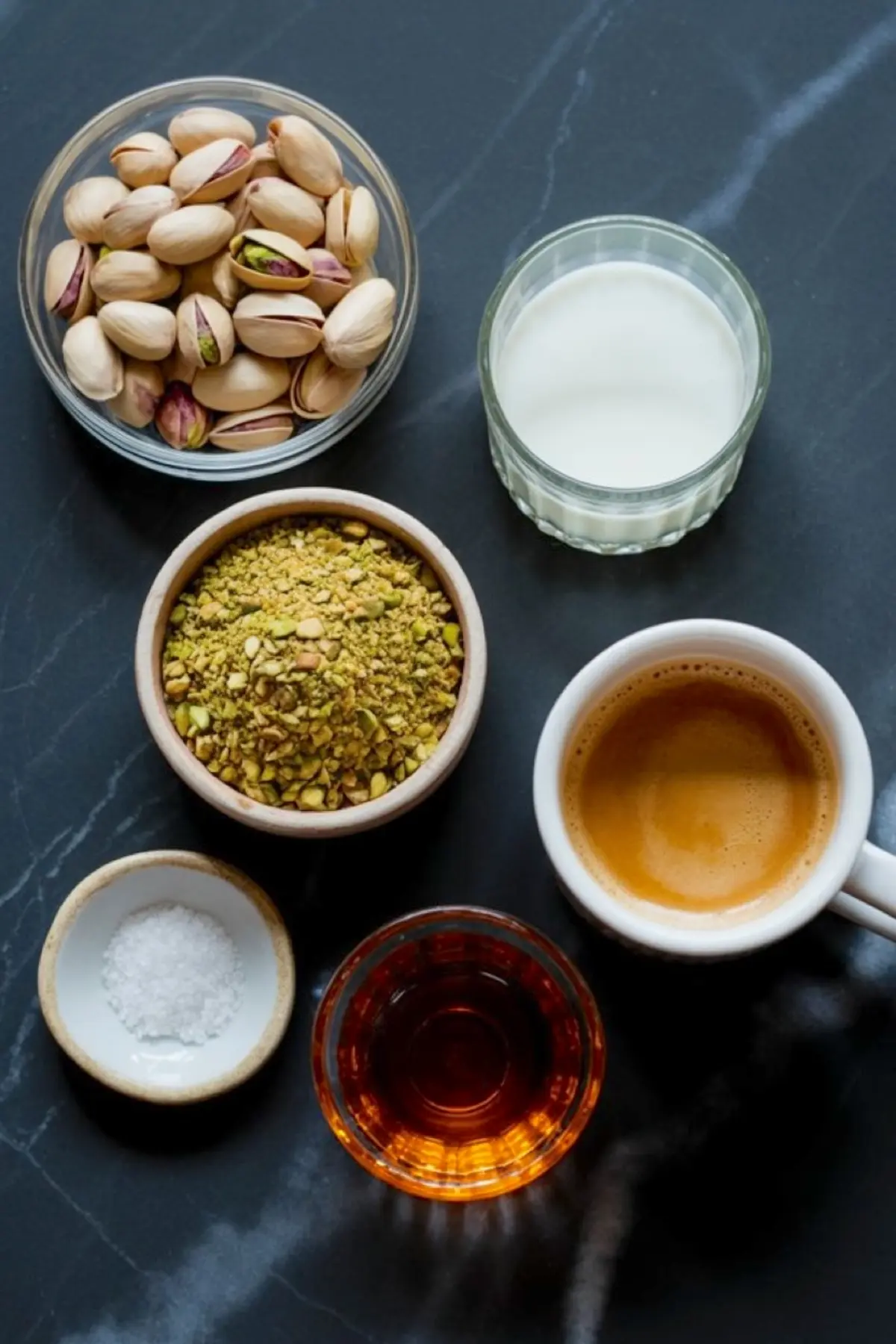 Flat lay of pistachio latte ingredients on a black marble surface, including whole pistachios, crushed pistachios, milk, espresso, maple syrup, and sea salt in small glass and ceramic bowls.