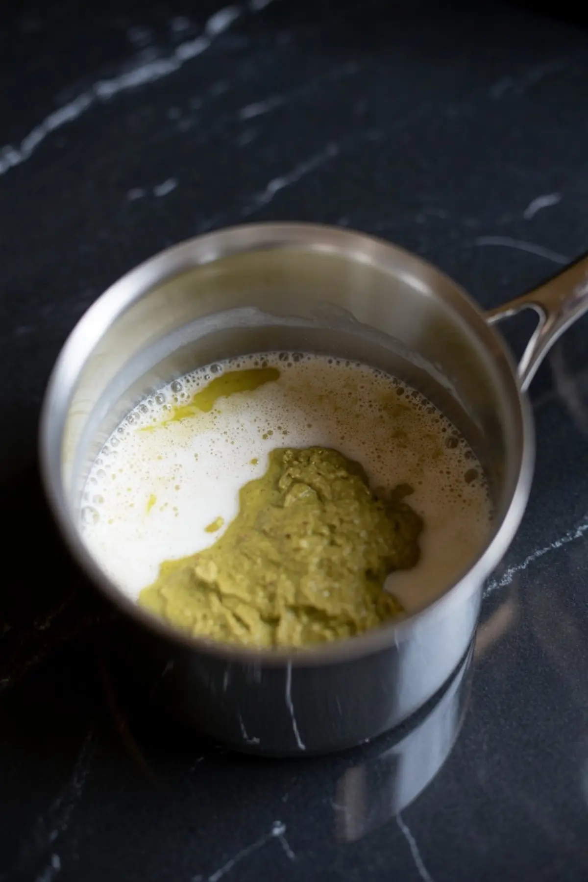 Ground pistachio paste mixed with milk in a stainless steel saucepan, beginning the process of making a pistachio milk base on a dark marble counter.