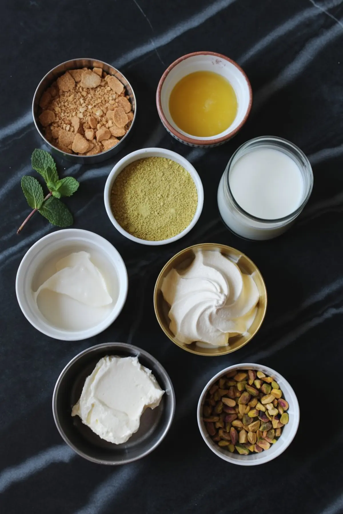 Overhead view of pistachio pie ingredients including graham cracker crumbs, melted butter, milk, pistachio pudding mix, whipped topping, Greek yogurt, cream cheese, and chopped pistachios arranged neatly on a dark countertop with fresh mint.