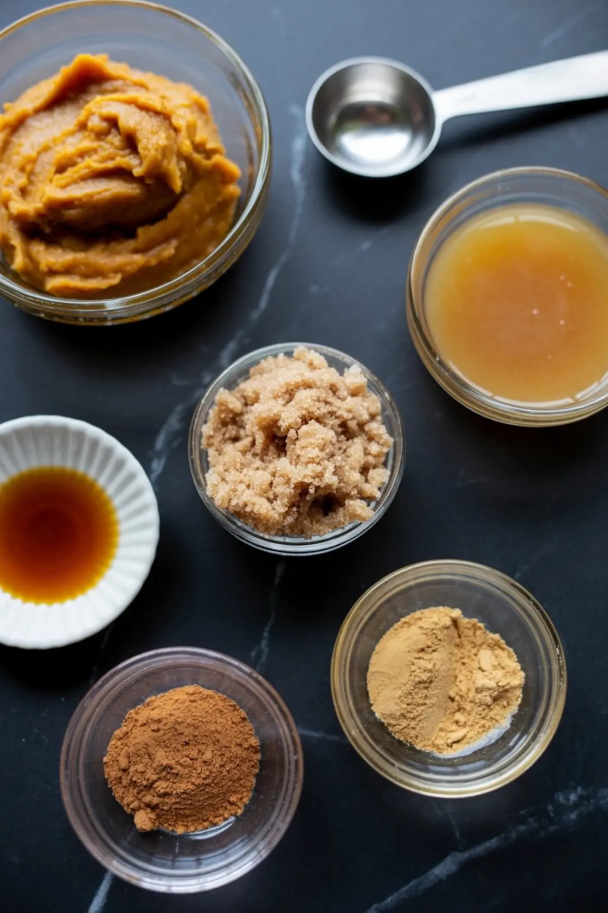 Overhead view of pumpkin butter ingredients on a dark surface, including pumpkin puree, apple cider, brown sugar, vanilla extract, ground ginger, ground cinnamon, and a measuring spoon.