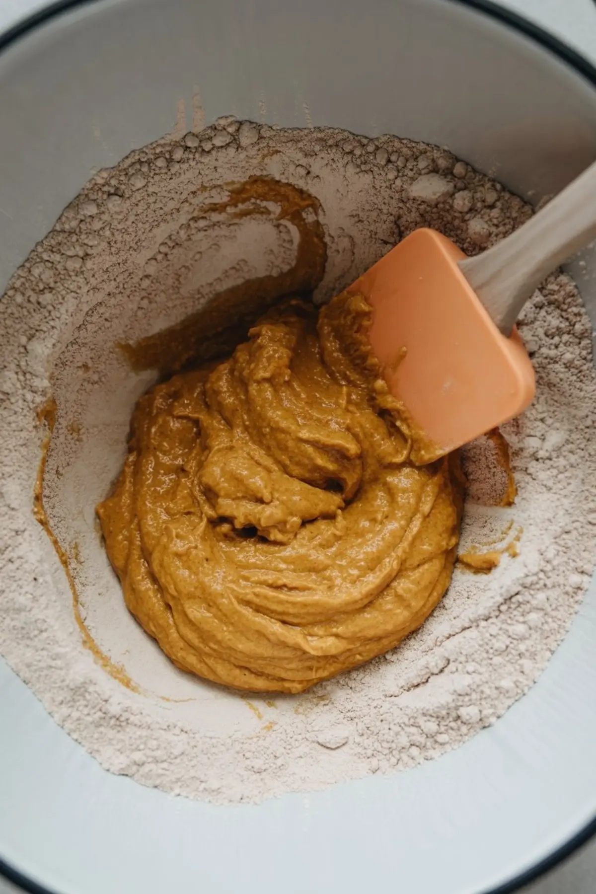 Pumpkin cake batter being folded with a spatula in a mixing bowl filled with flour, creating a smooth and creamy texture.