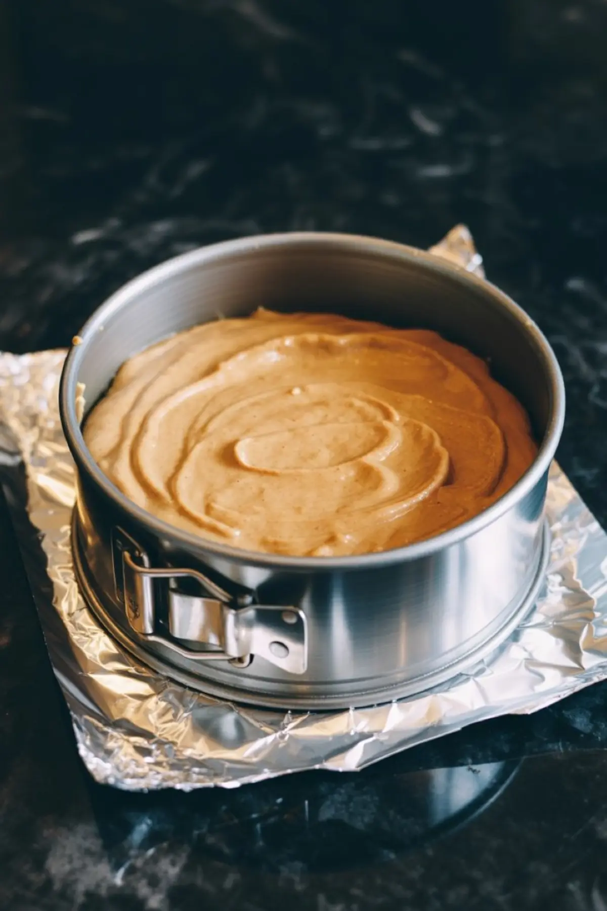 Pumpkin cheesecake batter poured into a foil-wrapped springform pan, ready for baking on a black marble counter.
