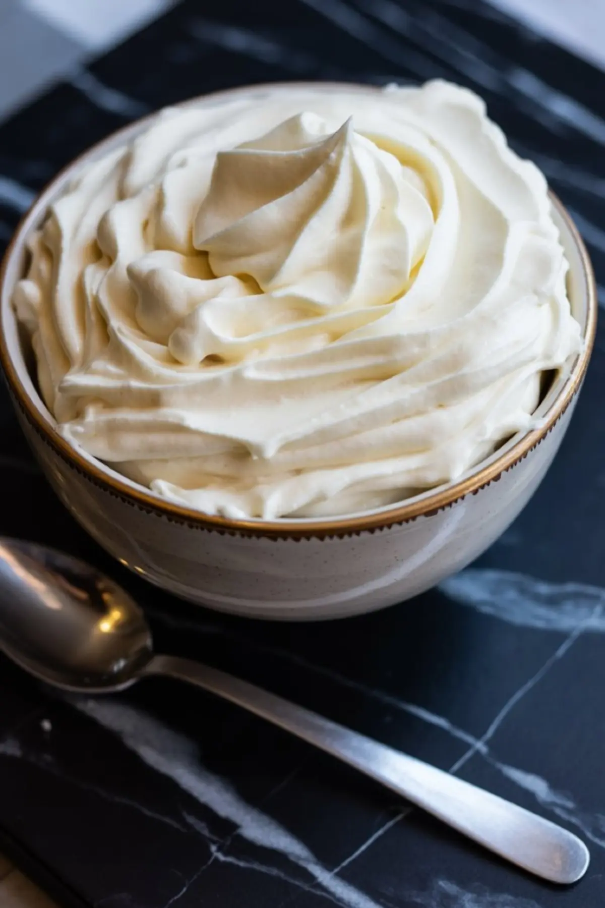 Bowl of whipped cream piped in soft swirls, placed on a black marble surface with a silver spoon beside it.