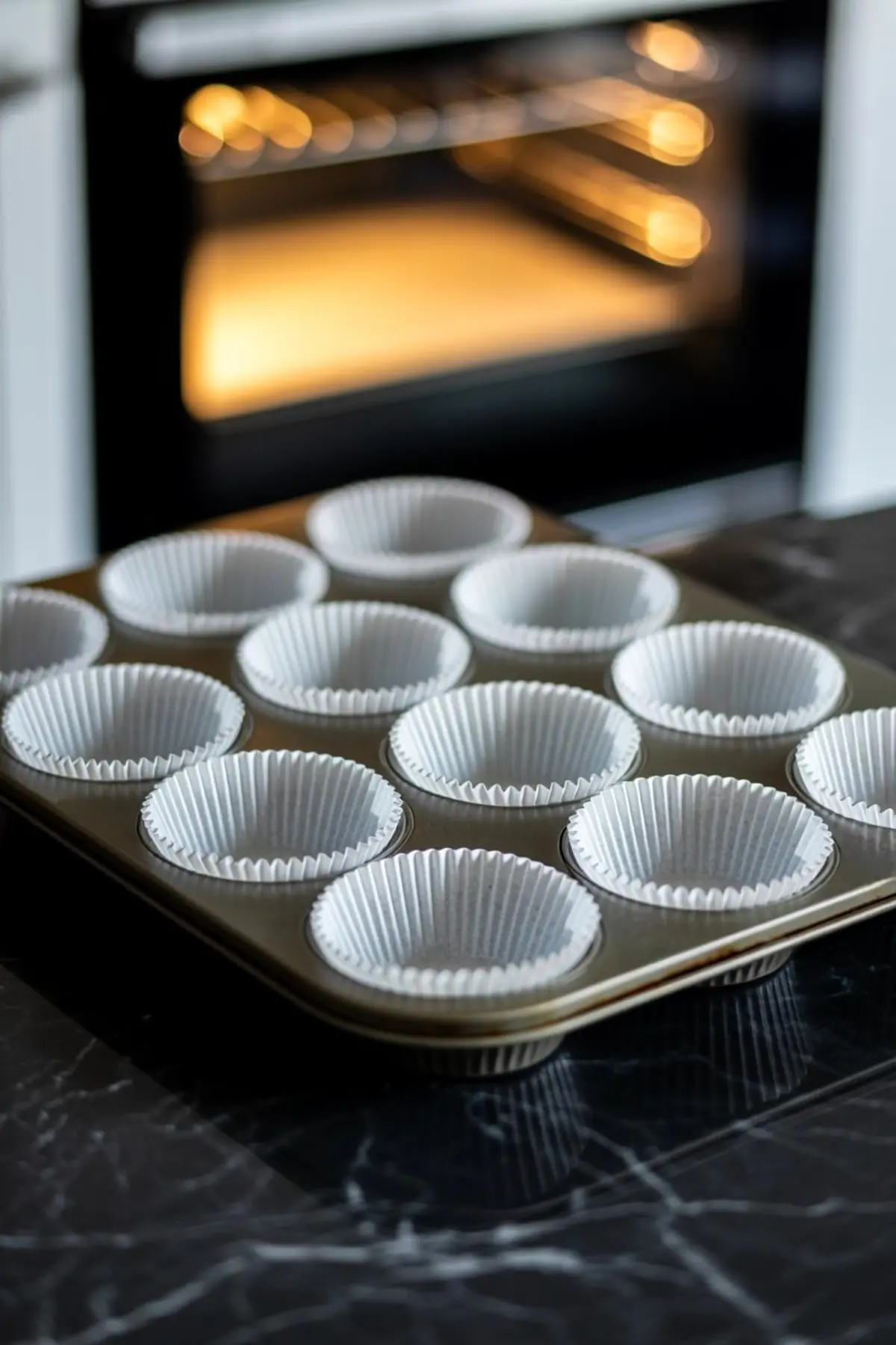 Metal cupcake tin lined with white paper liners set on a black marble counter, with an oven glowing in the background.