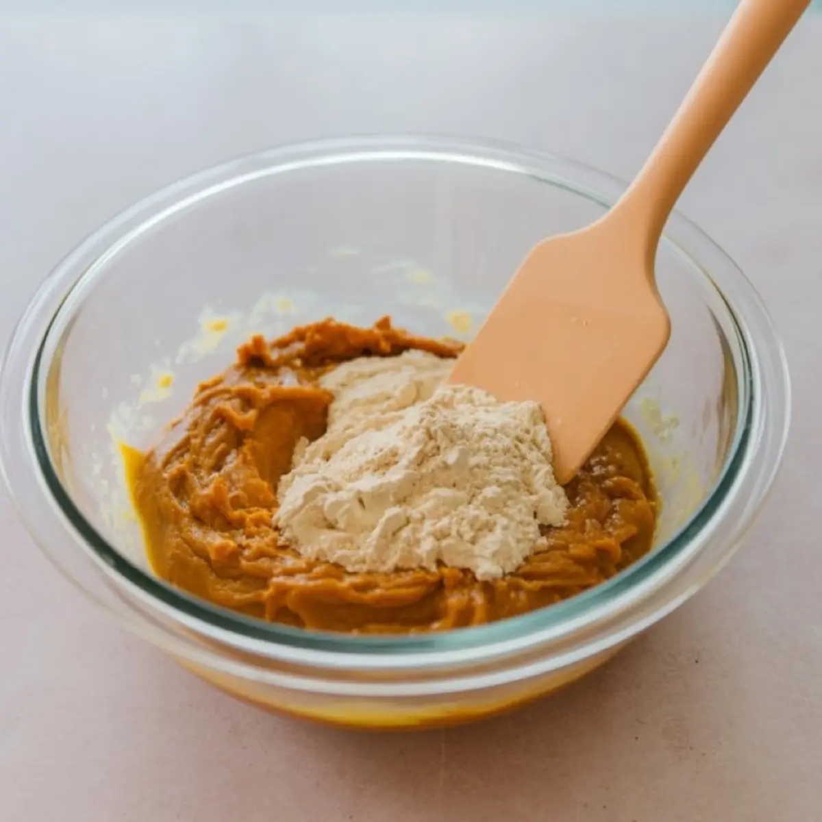 Pumpkin mixture and dry ingredients combined in a glass mixing bowl with a peach spatula, placed on a light pink surface.