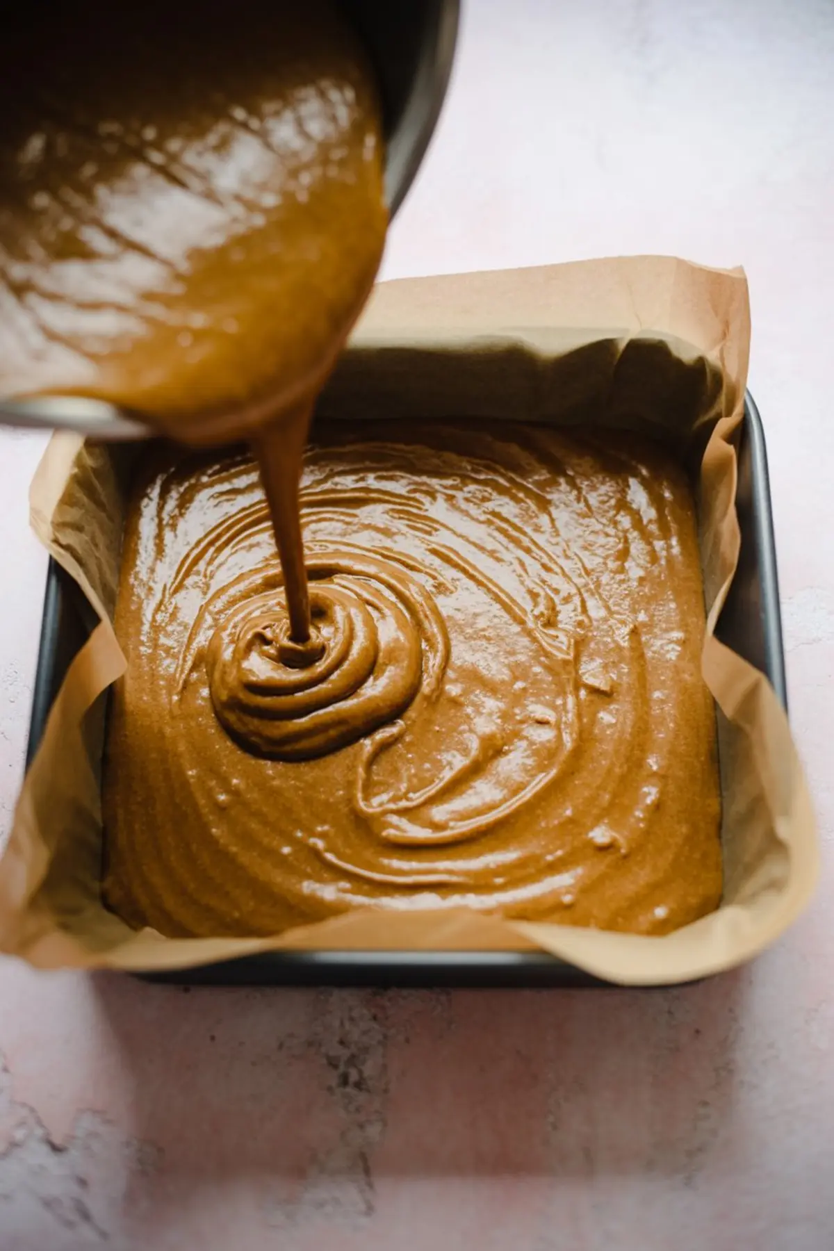 Warm pumpkin fudge mixture being poured from a saucepan into a parchment-lined square baking pan. The thick, glossy batter swirls as it fills the tray, showcasing the final stage before setting. A key step in making soft homemade fudge.