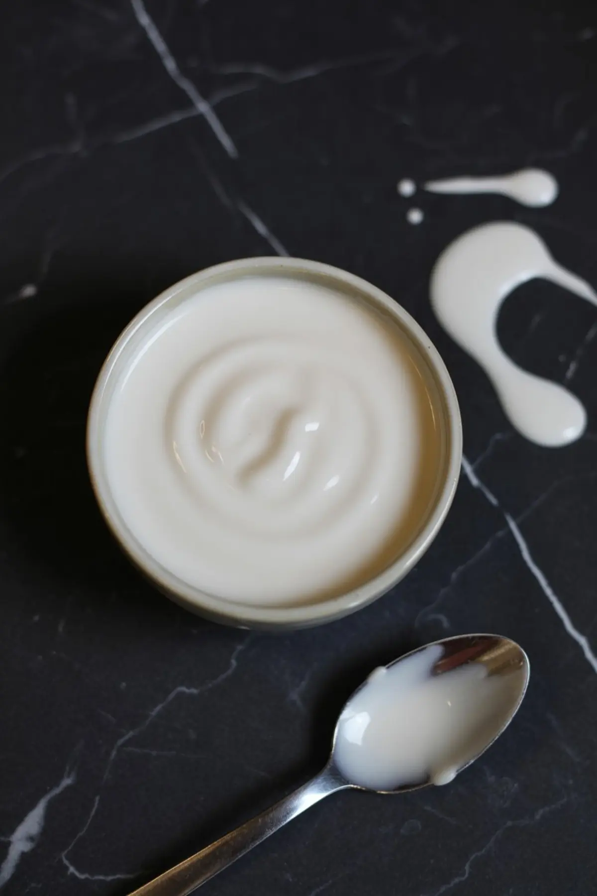Bowl of creamy Greek yogurt with a spoon and small splashes on a black marble surface, showing texture and consistency.