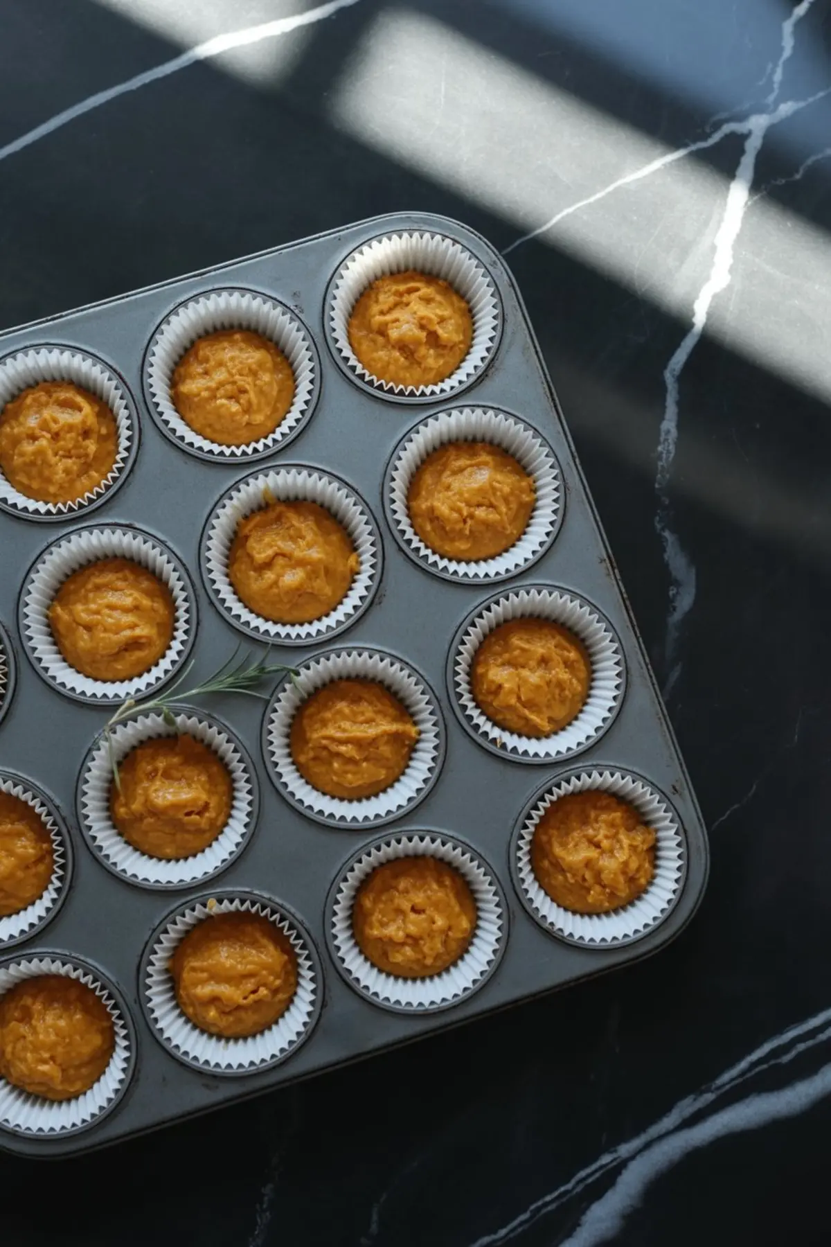 Overhead shot of a muffin tray filled with raw pumpkin muffin batter in paper liners, placed on a black marble surface with a sprig of rosemary.