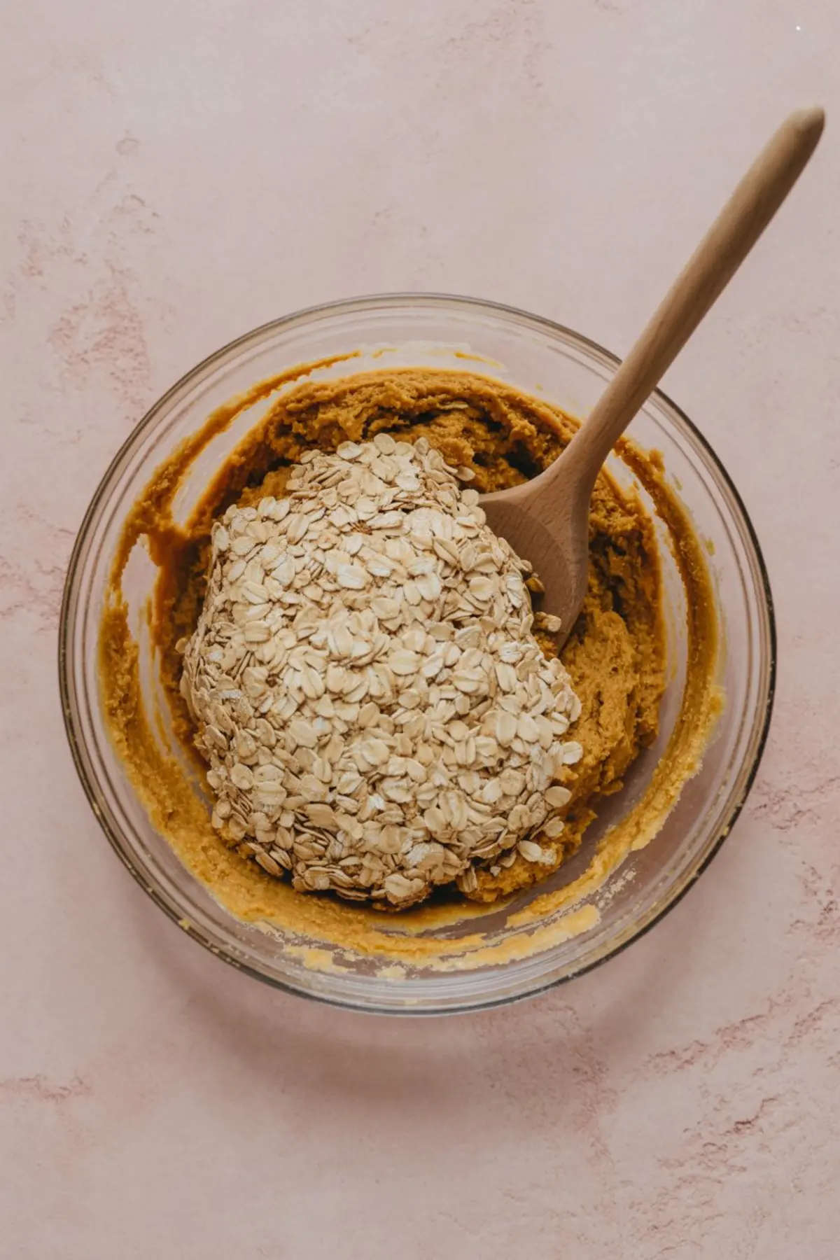 Mixing bowl filled with pumpkin cookie dough partially blended with rolled oats, with a wooden spoon resting in the bowl, placed on a pink textured surface.