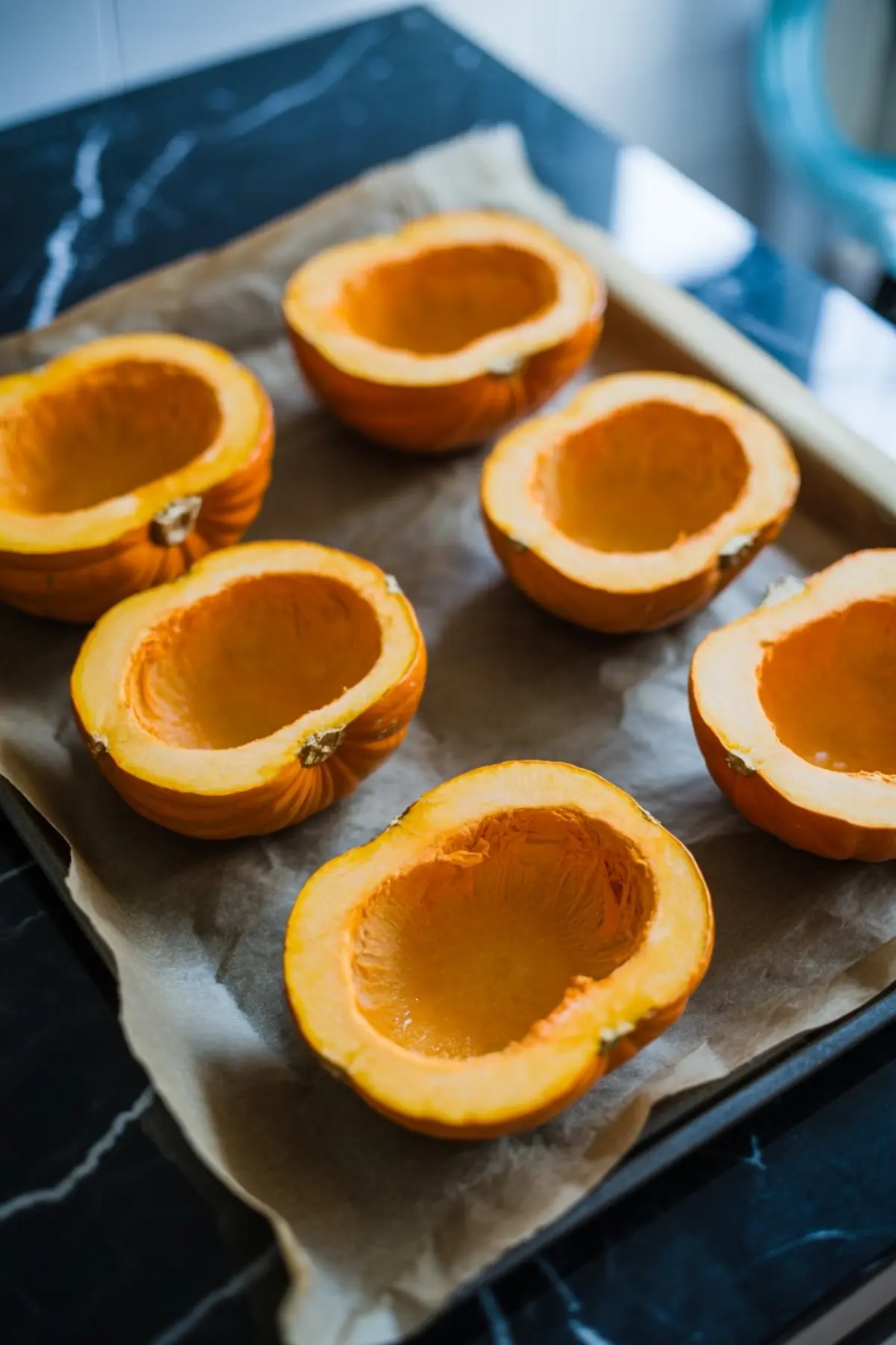 Baking tray lined with parchment paper holding six halved pumpkins with seeds removed, ready for roasting.