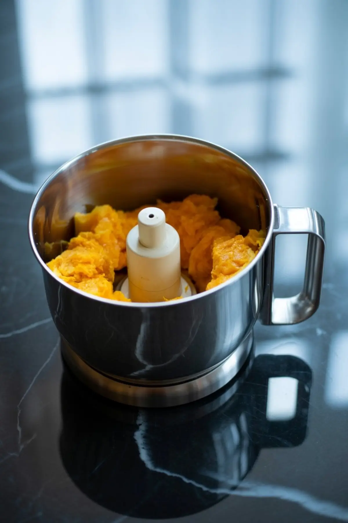 Stainless steel food processor bowl filled with chunks of cooked pumpkin on a dark marble countertop.