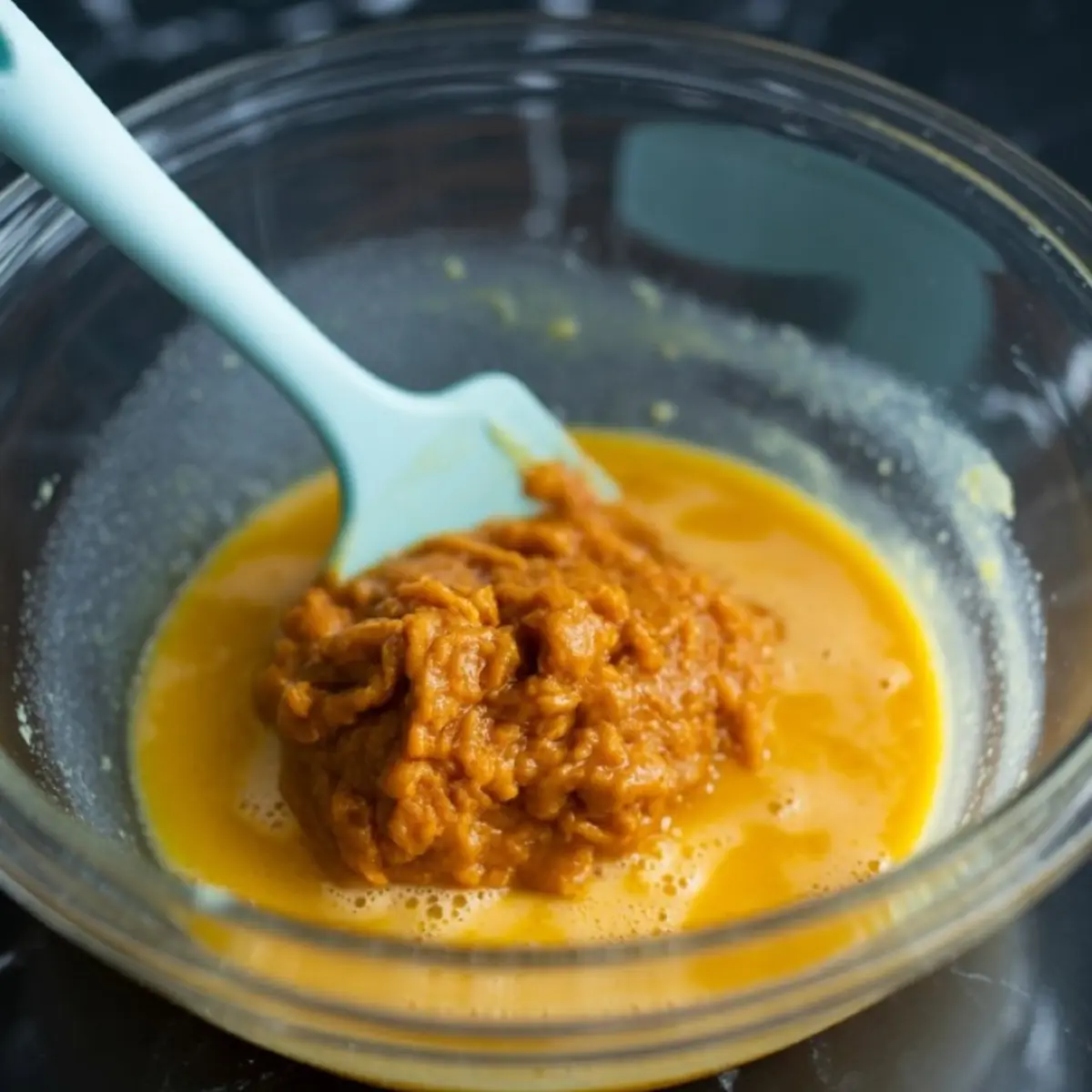 Pumpkin puree mixed with wet ingredients in a clear glass bowl using a light blue spatula.