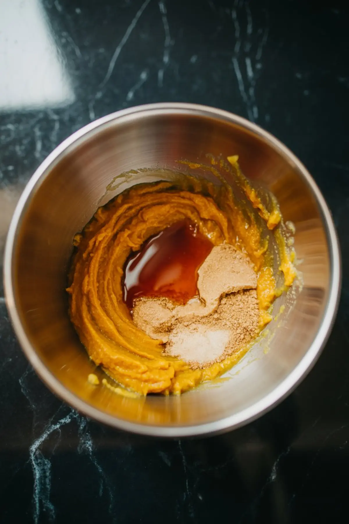 Mixing bowl with smooth pumpkin purée partially blended with sugar, vanilla extract, and spices, sitting on a black countertop.