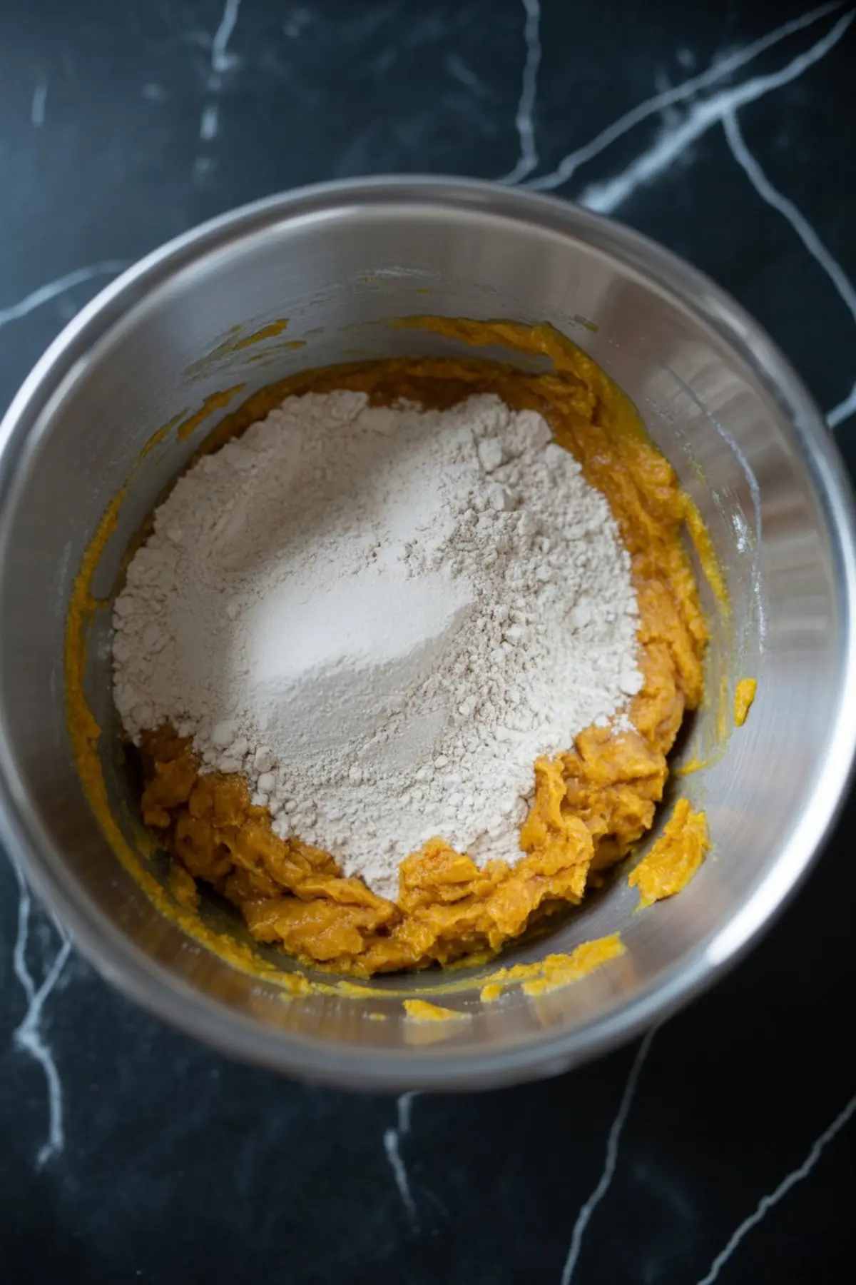 Metal mixing bowl showing thick pumpkin batter with flour added on top, ready to be blended for cookie dough.