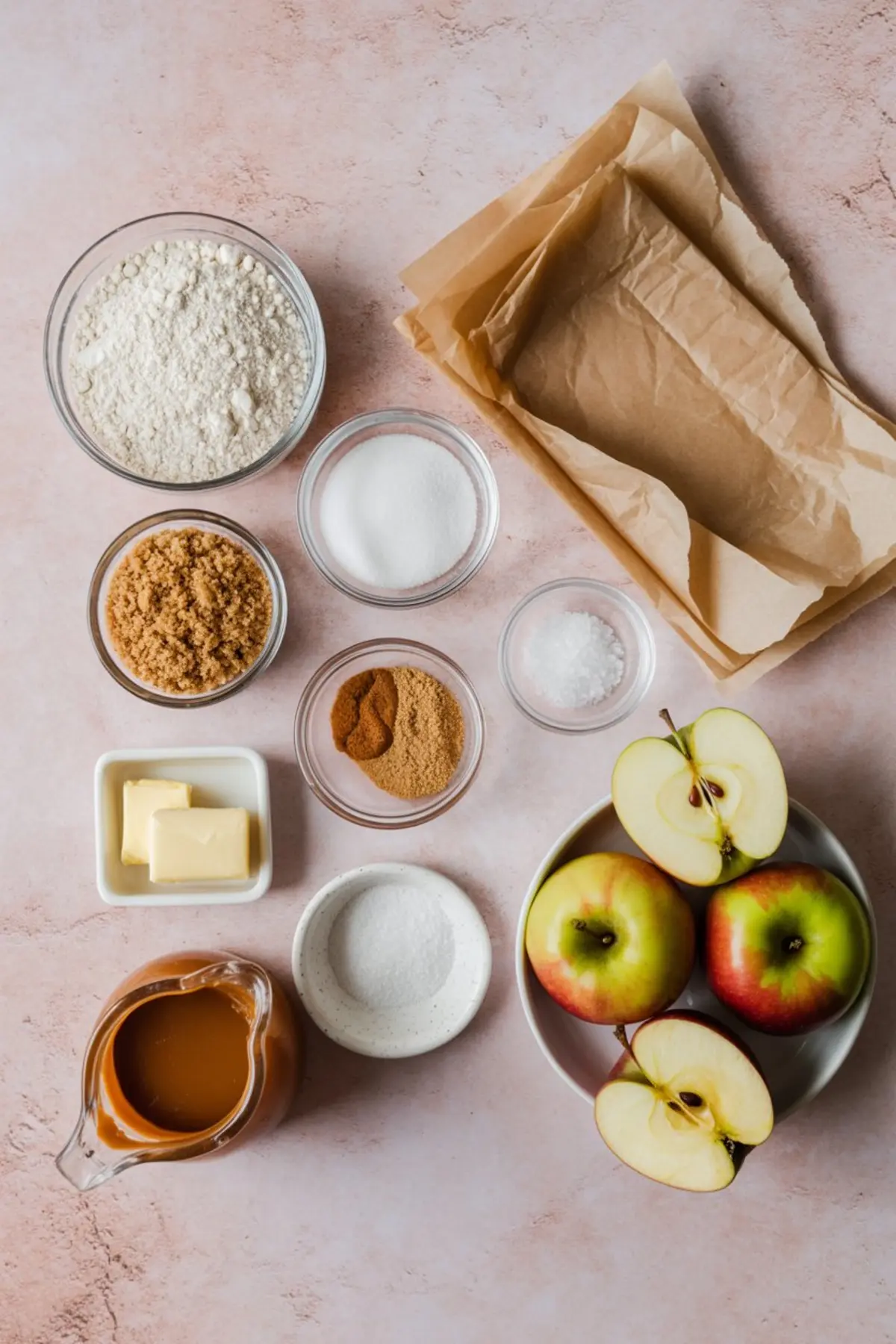 Flat lay of baking ingredients for salted caramel apple pie bars, including flour, sugars, butter, spices, apples, salt, and caramel sauce, all arranged on a pink backdrop.