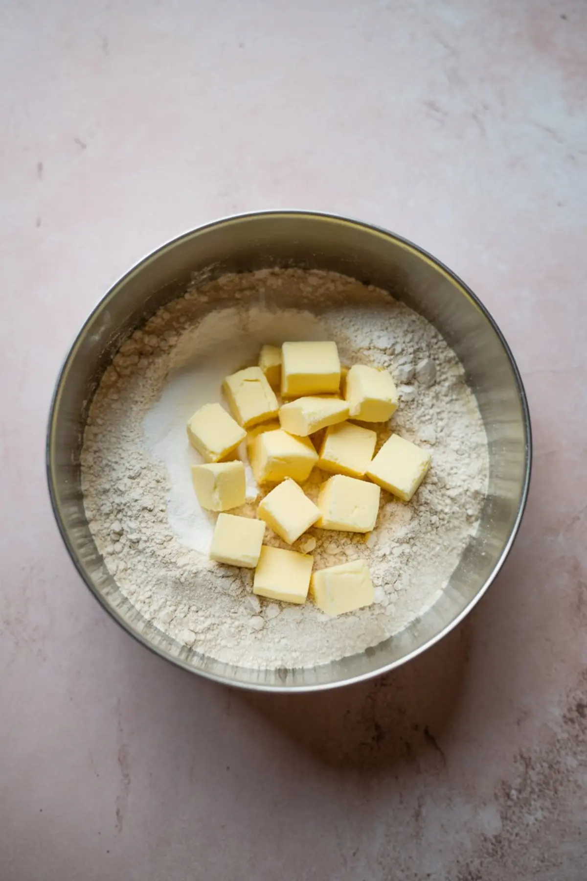 Metal bowl filled with flour and sugar topped with cubed cold butter, set on a pale pink surface for making a buttery shortbread crust.