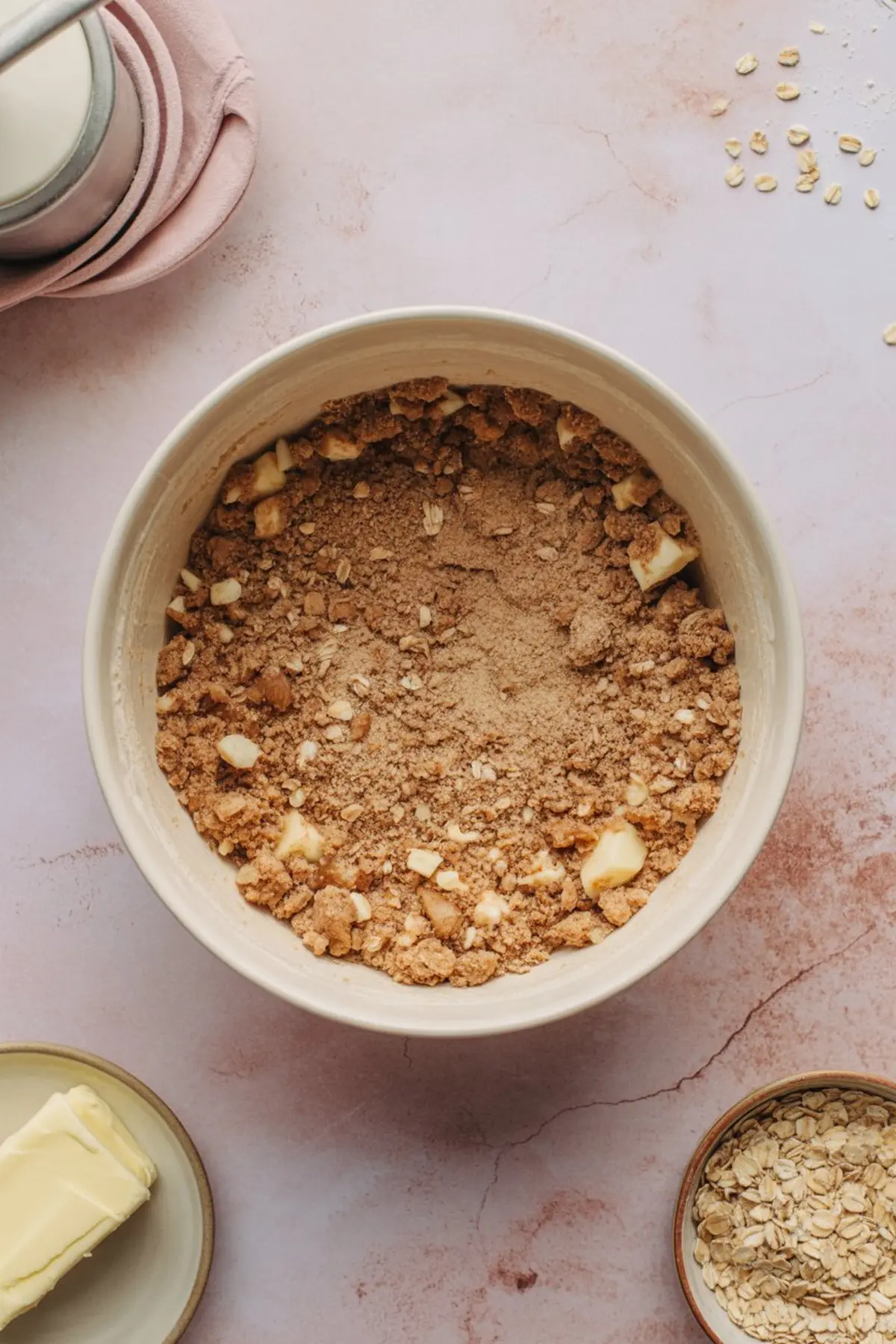 Mixing bowl containing oat crumble mixture with brown sugar, butter, and flour chunks, surrounded by additional ingredients on a light pink countertop.