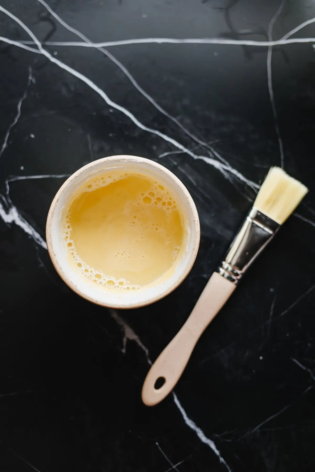 Beaten egg wash in a ceramic bowl with a pastry brush on black marble counter, used for brushing pie crusts.
